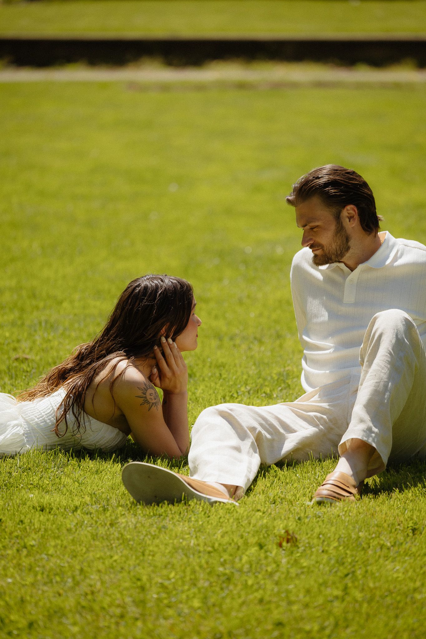 Couple in white outfits sitting on green grass in a park, sharing an intimate conversation on a sunny day.