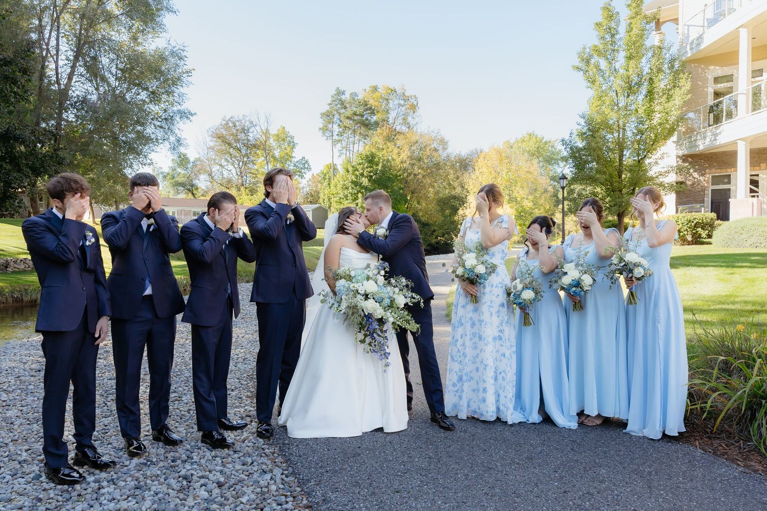 Wedding party poses outdoors, groomsmen covering eyes as bride and groom kiss, bridesmaids in light blue gowns.