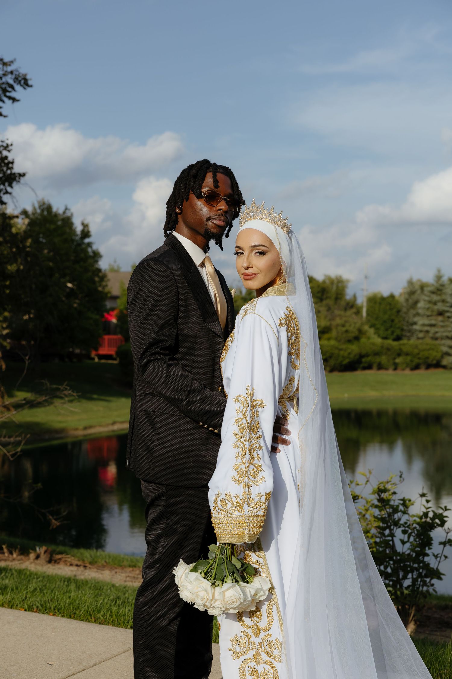 Interracial couple in wedding attire pose outdoors, groom in black suit, bride in white and gold hijab gown with bouquet.