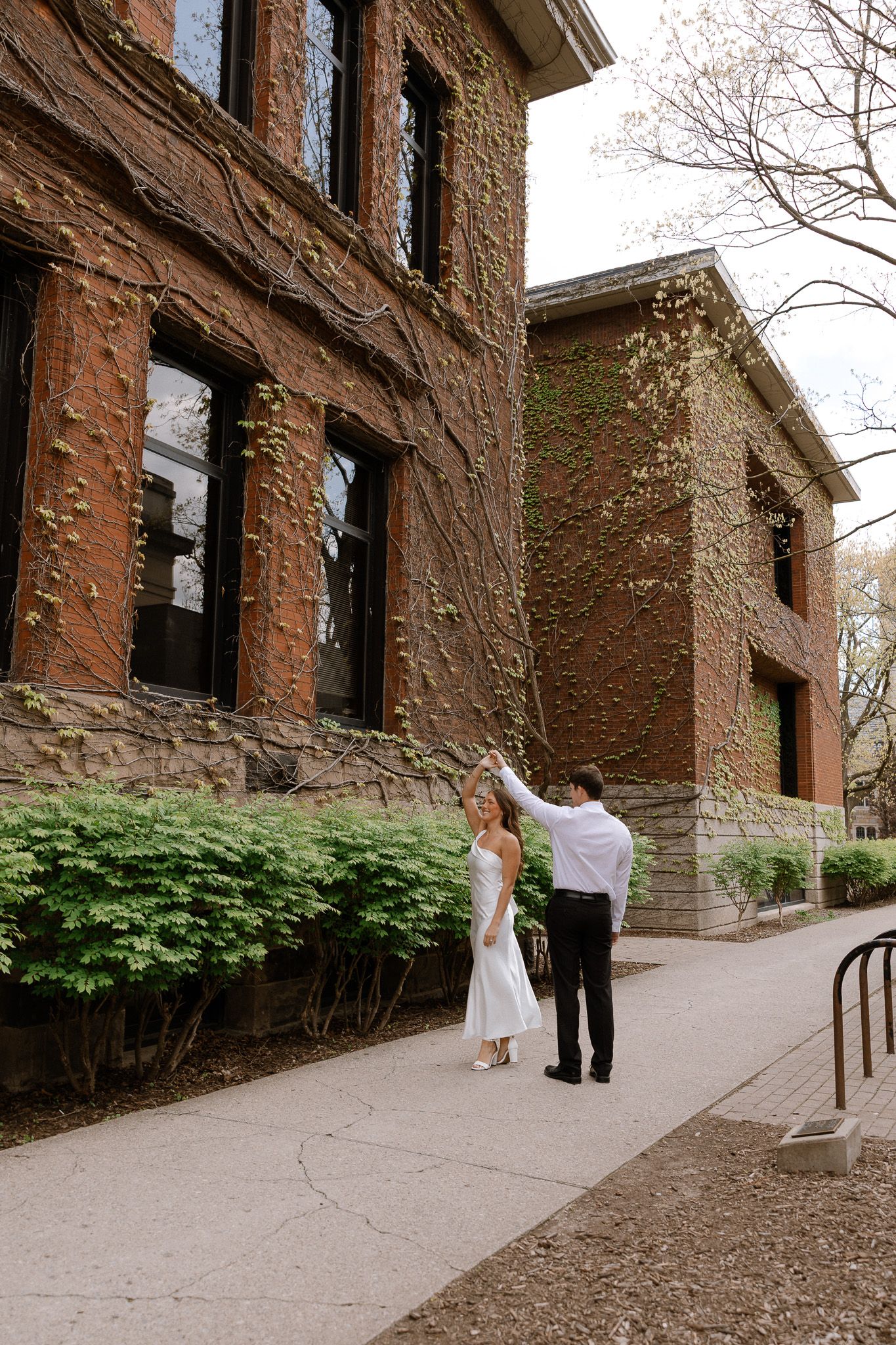 Couple in wedding attire dancing outside ivy-covered brick building on a sunny day.
