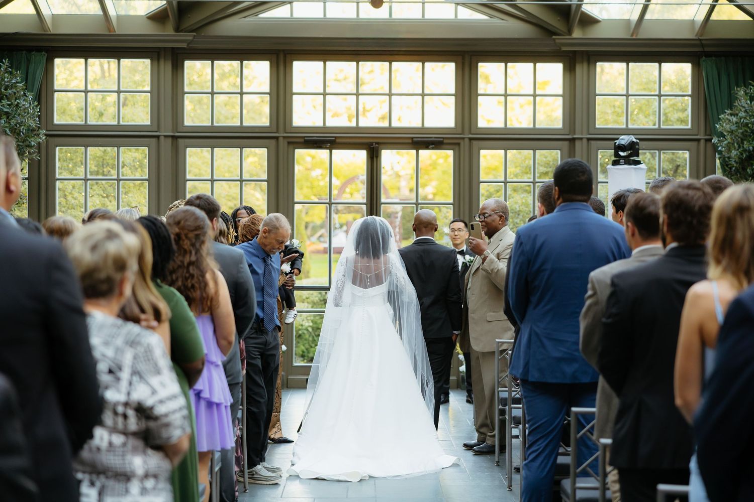 Bride walks down the aisle in a white gown and veil at an elegant indoor wedding ceremony with guests standing.