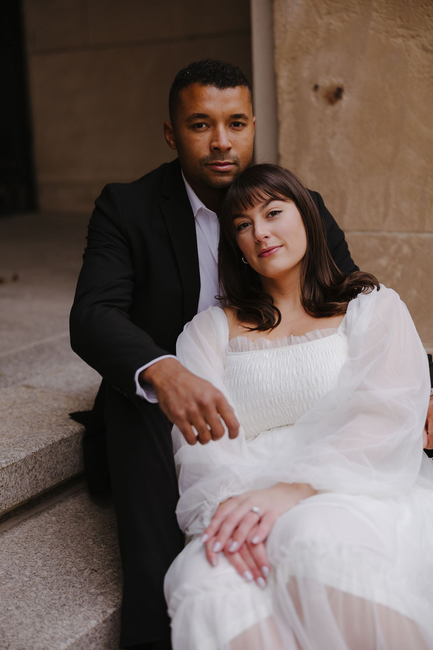 A couple in formal attire poses intimately against a stone wall backdrop.