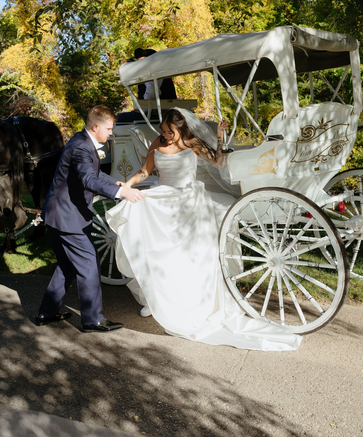 Bride in white gown steps out of elegant white horse-drawn carriage assisted by groom in navy suit outdoors.