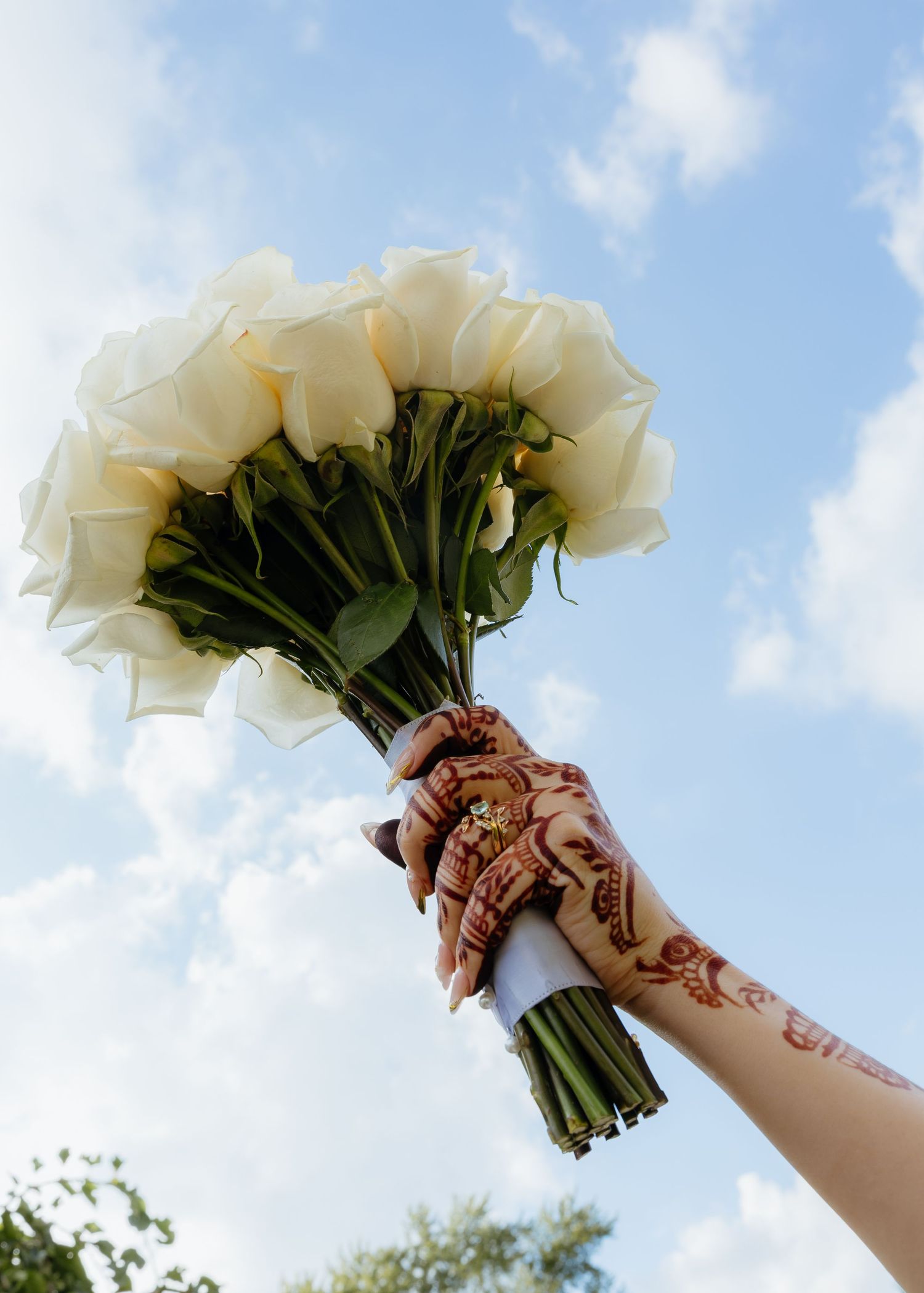 A henna-adorned hand holds a bouquet of white roses against a blue sky at a wedding ceremony.