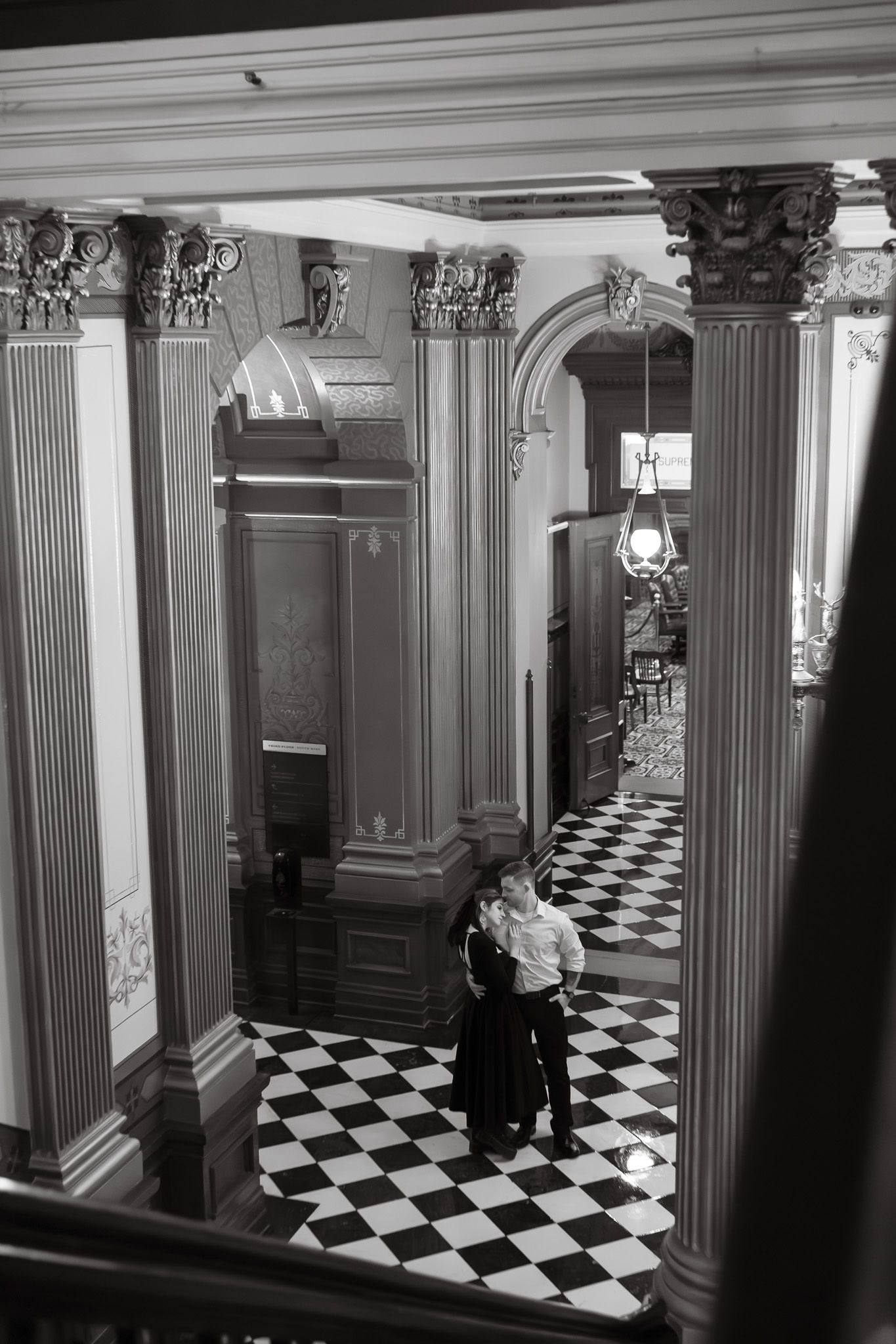 Black and white aerial view of couple embracing in grand hall with checkered floors and ornate columns.