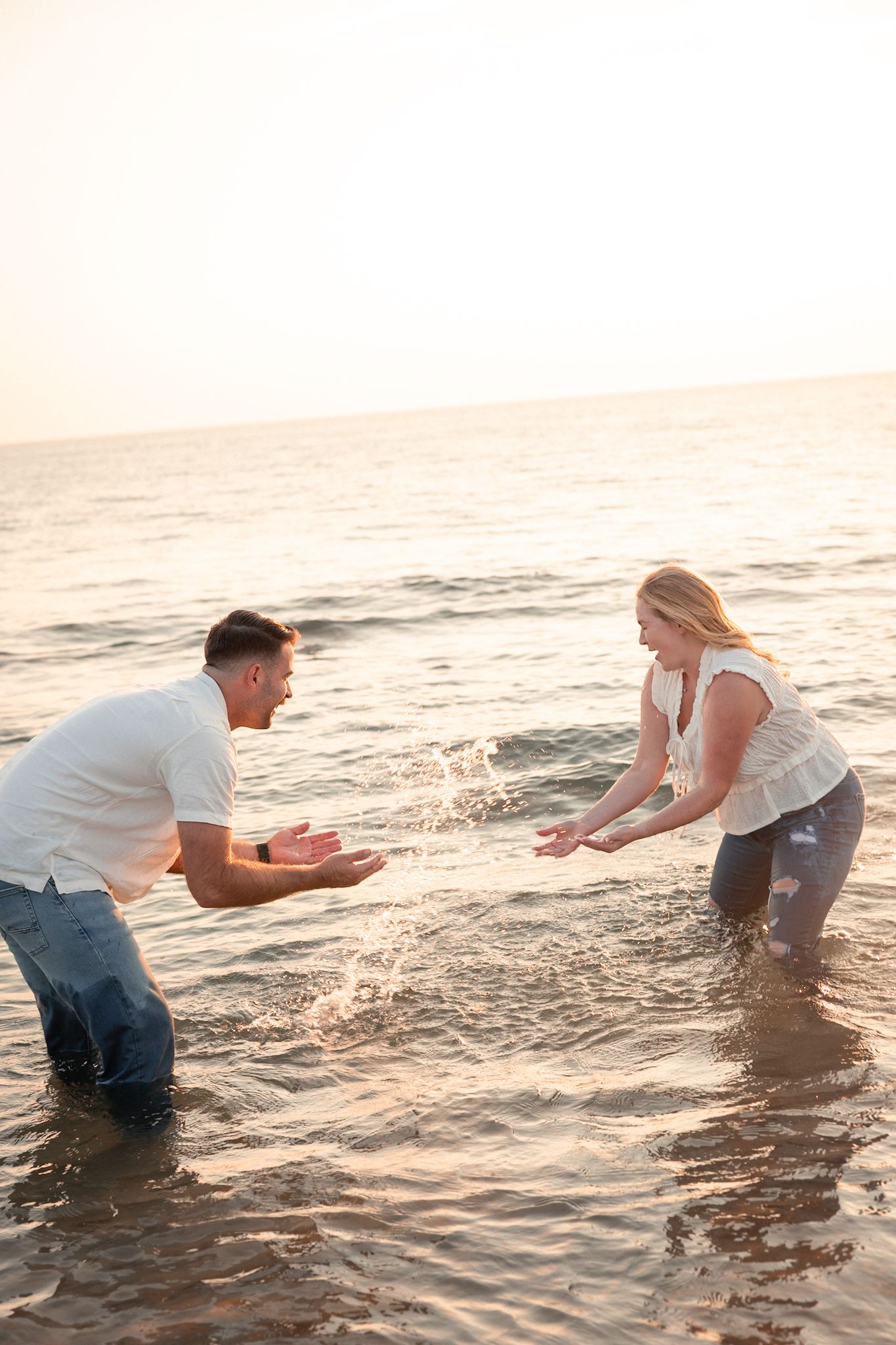 Playful couple splashes in ocean waves during sunset beach photoshoot.