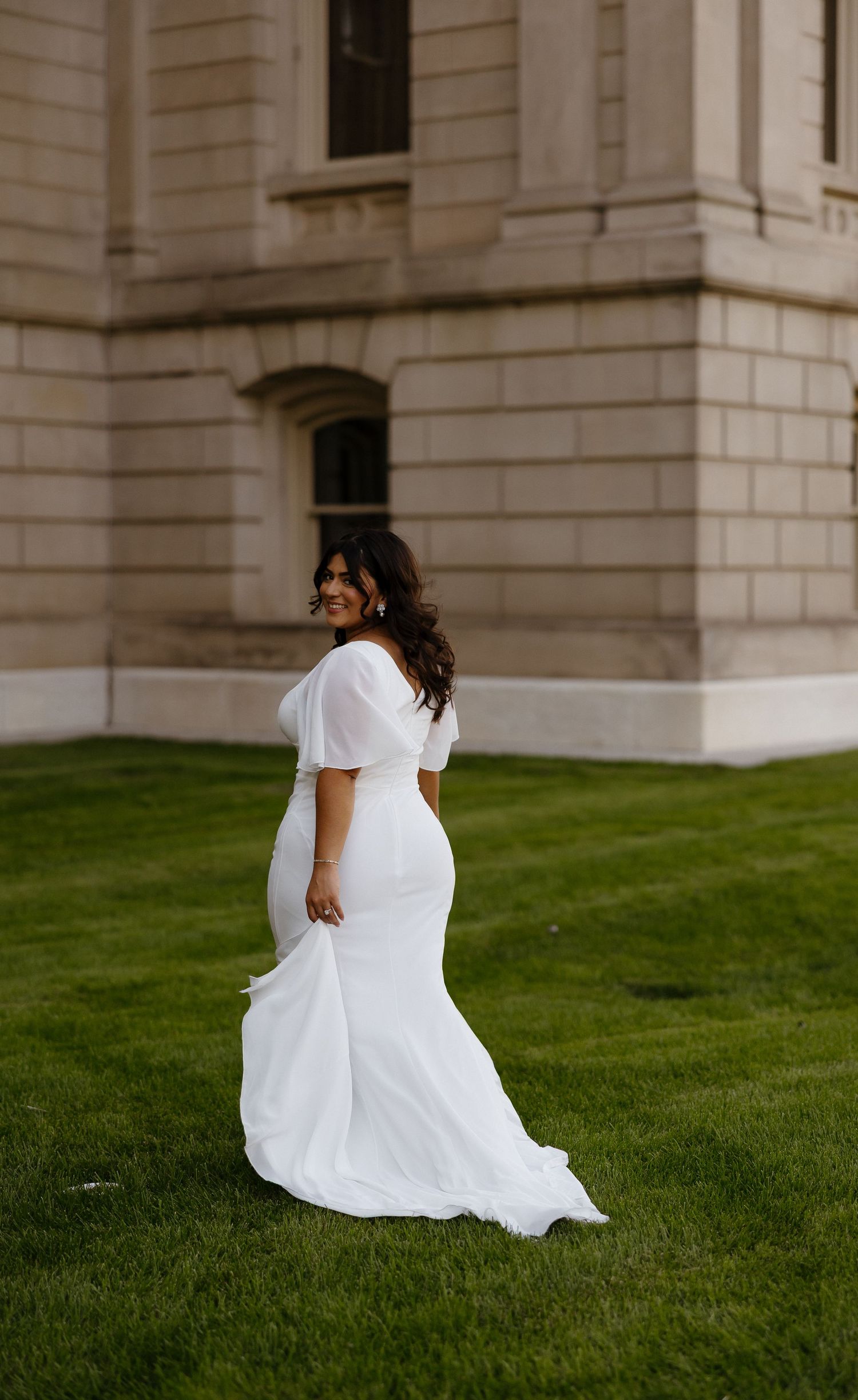 Person in flowing white dress walks gracefully across manicured lawn by stone building.