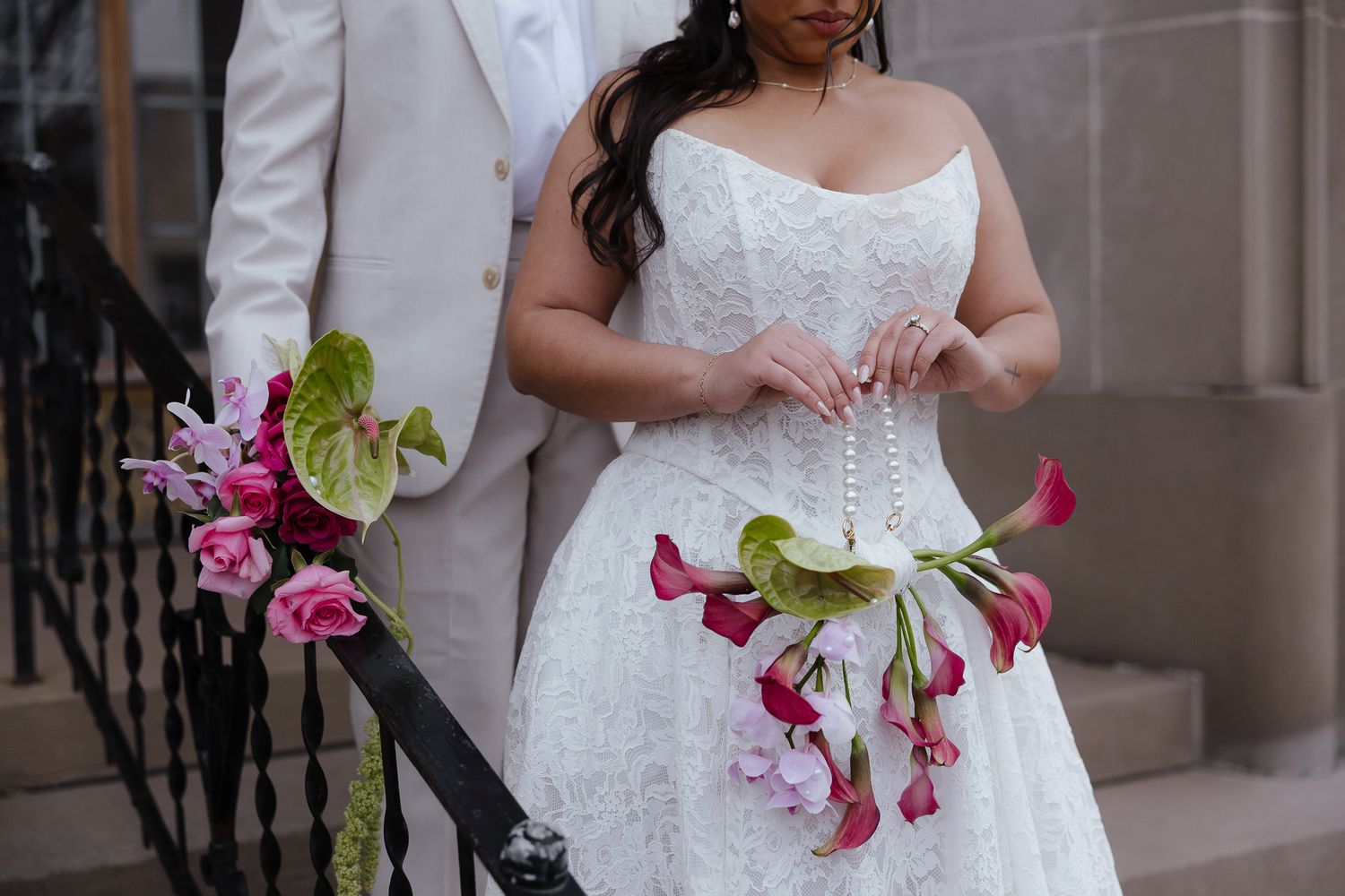 A bride in a white lace wedding dress holds a vibrant bouquet of pink calla lilies with green accents.