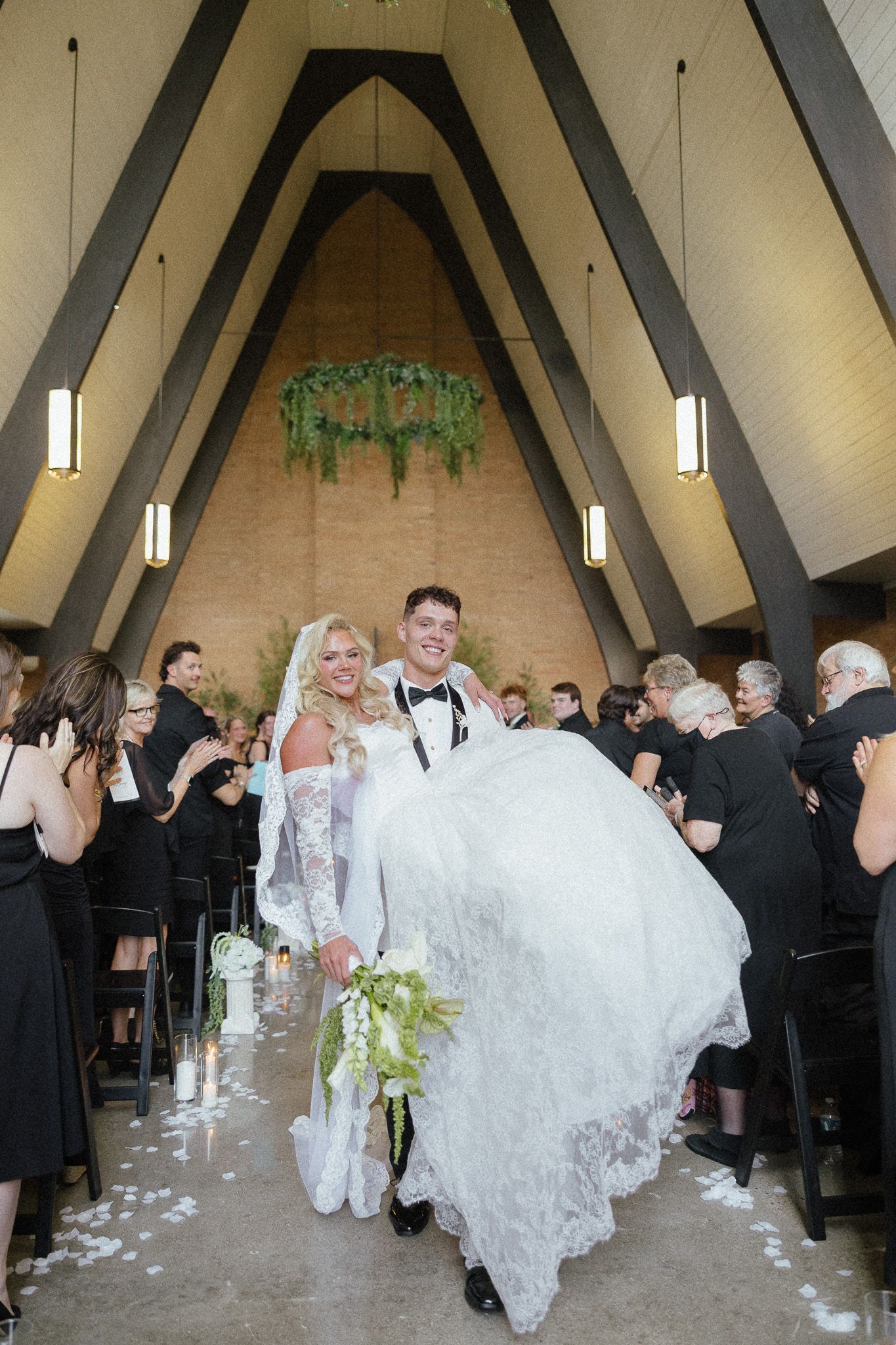 Groom carries bride down chapel aisle after wedding ceremony, guests in black attire celebrate around them.