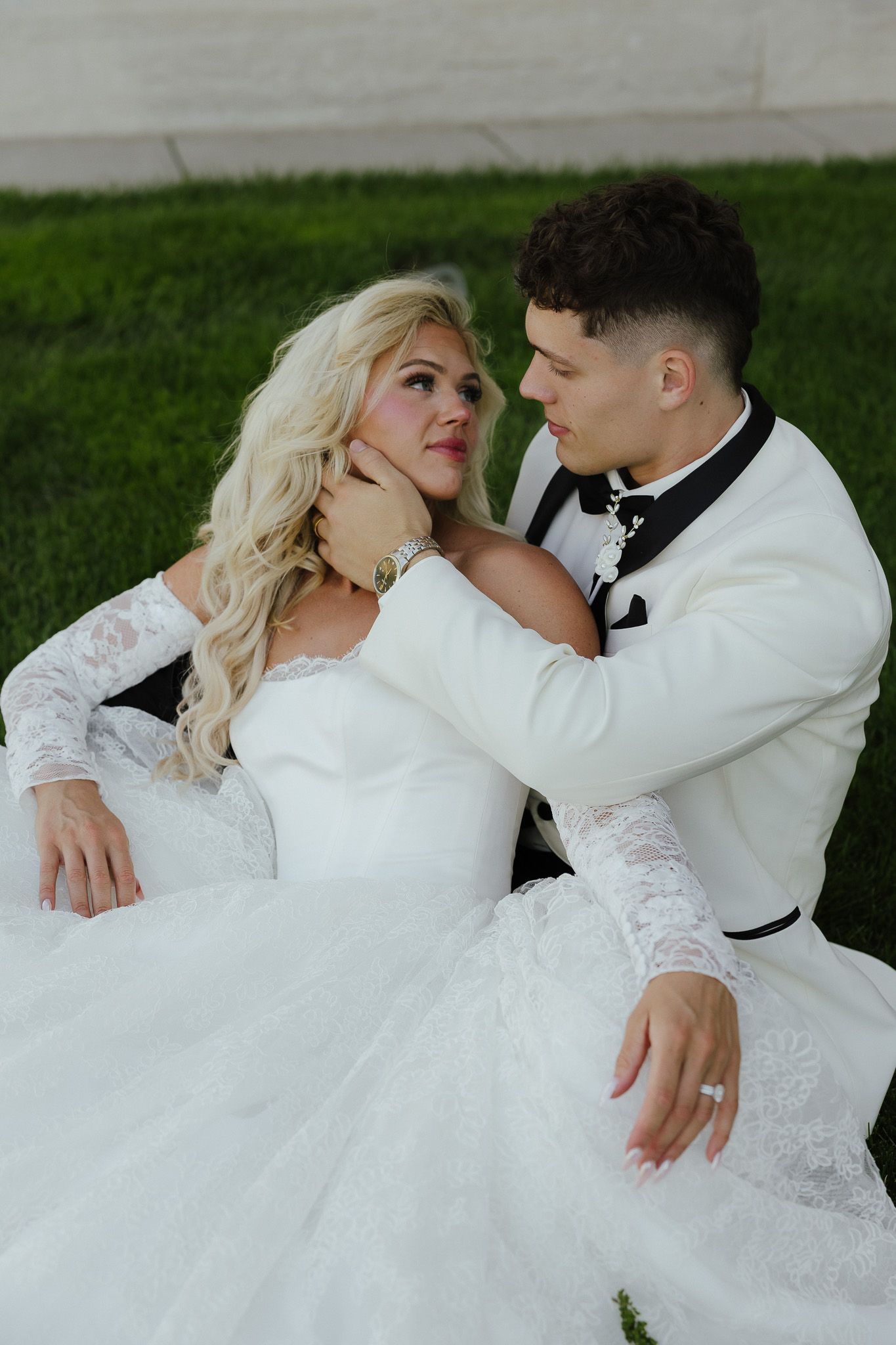 Bride and groom sitting on grass, groom in white tuxedo tenderly holding bride's face as they gaze at each other.