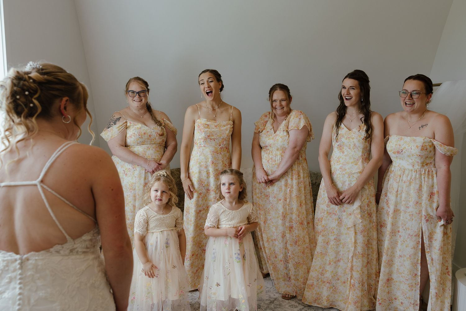 Bridesmaids in floral gowns react with joy seeing the bride, with two flower girls in white dresses standing in front.