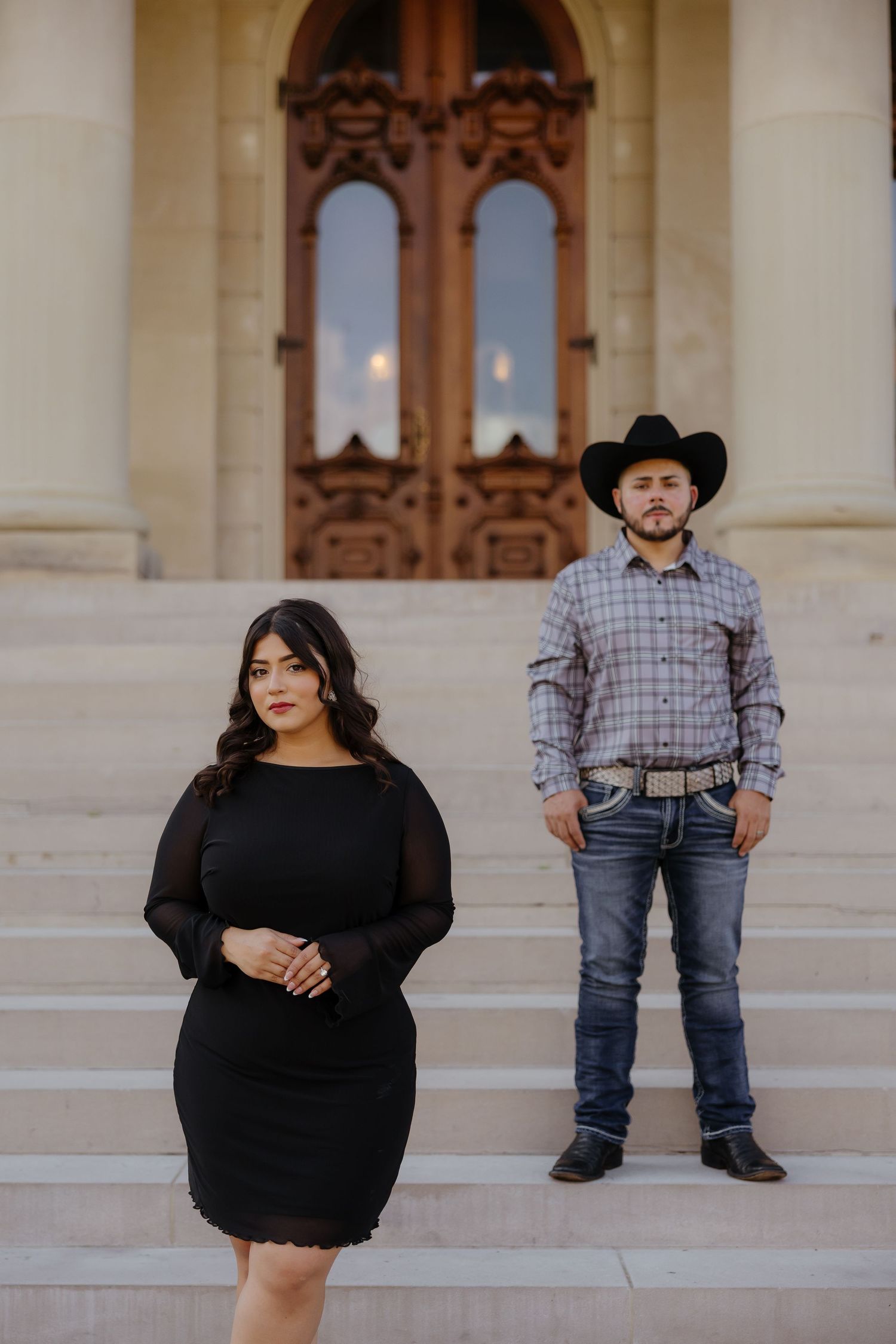 Stylish couple in western attire pose for engagement photos on stone steps of a grand building.