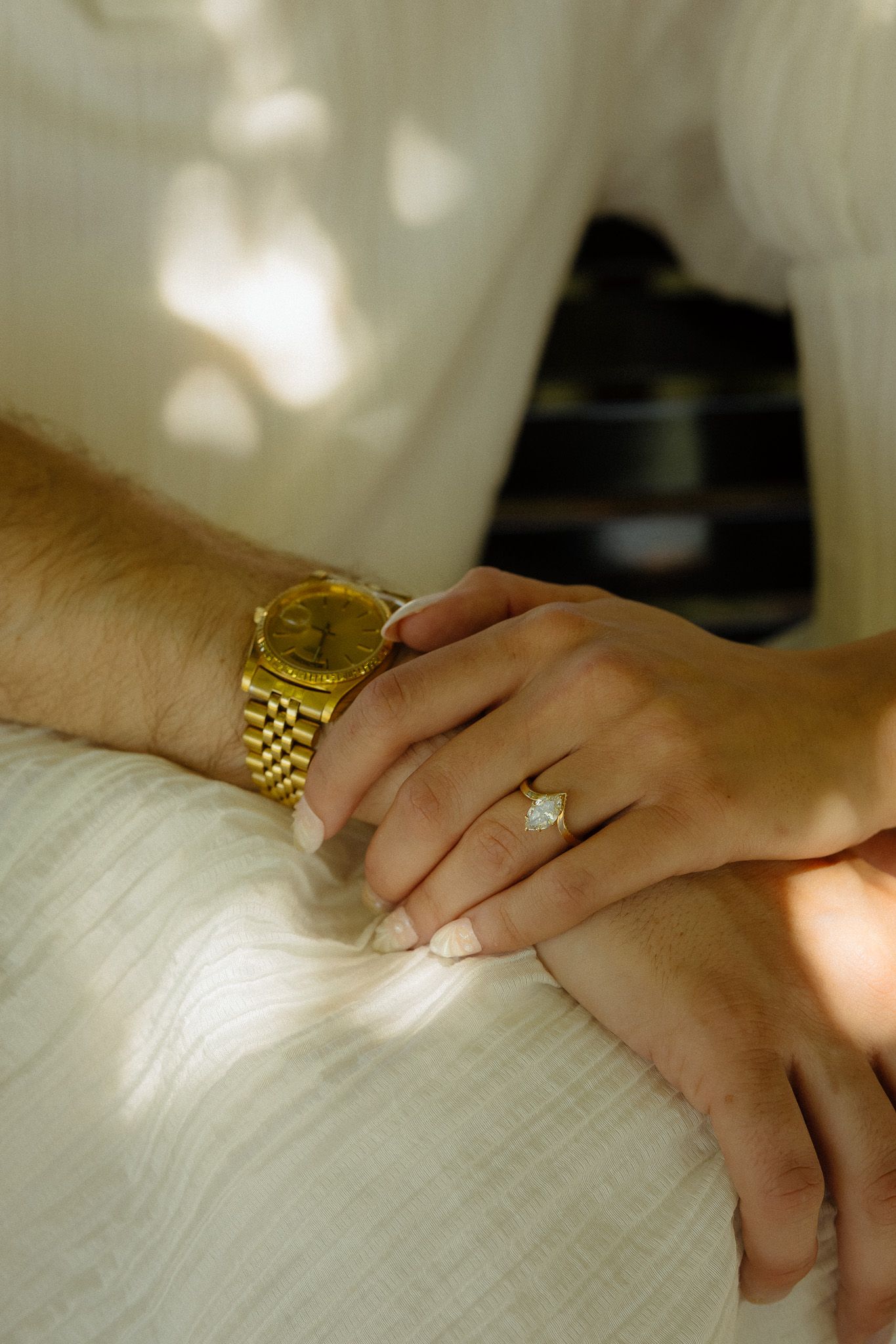 Two hands resting together, one wearing a gold Rolex watch and the other adorned with a diamond engagement ring.