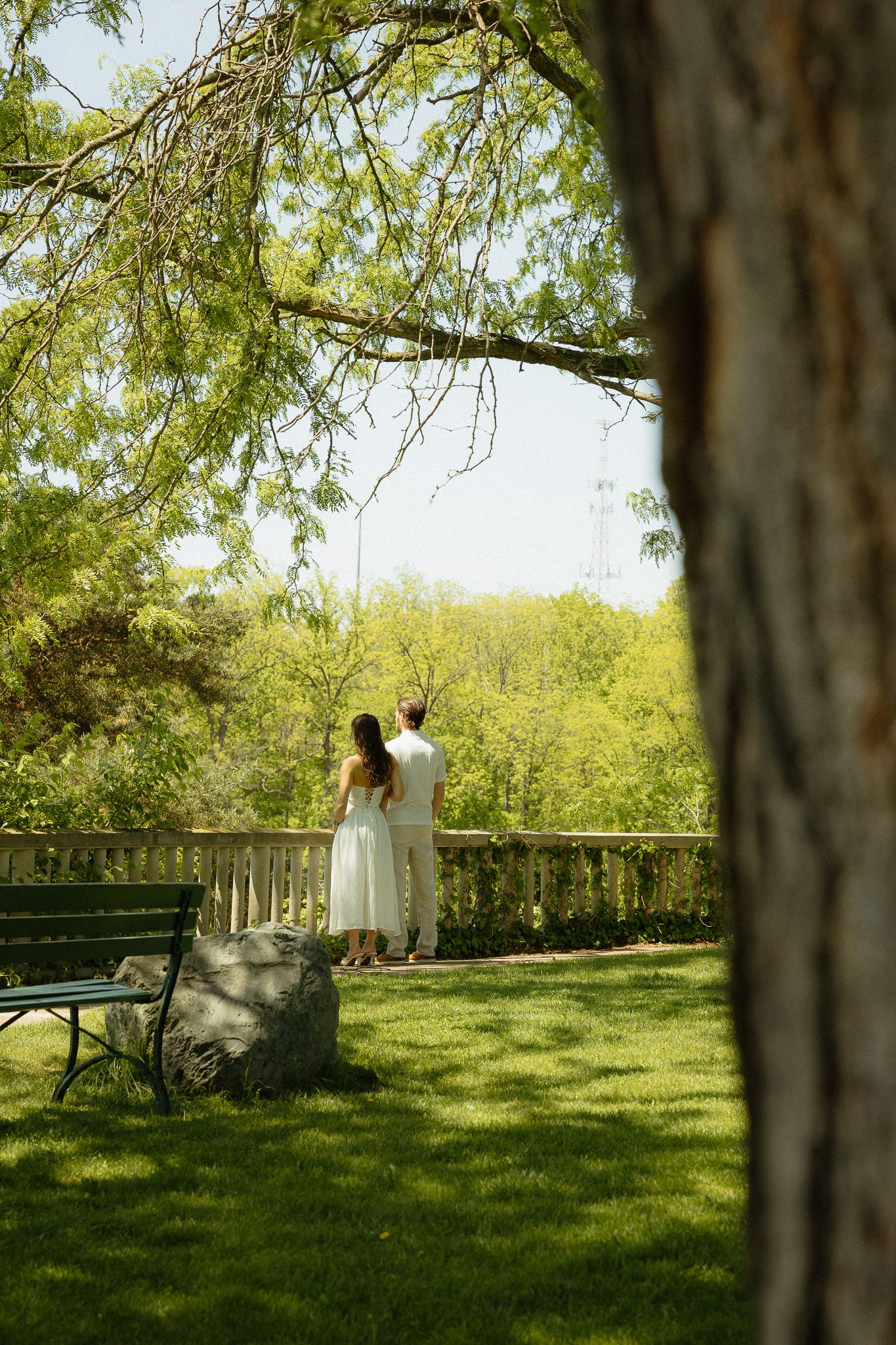 A couple in white attire stands together on a garden terrace, surrounded by lush green trees and manicured lawn.