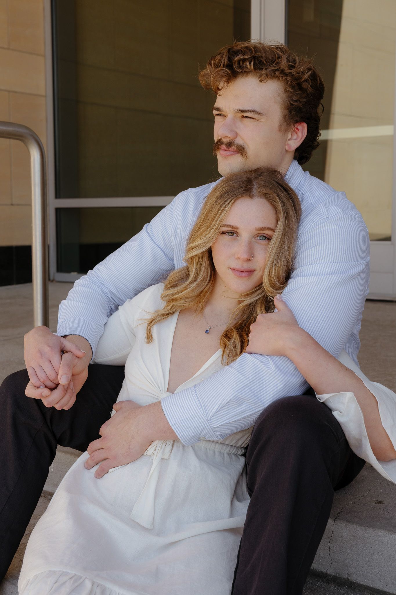 A young couple sits on steps, man in blue shirt embracing woman in white dress, both gazing thoughtfully into the distance.