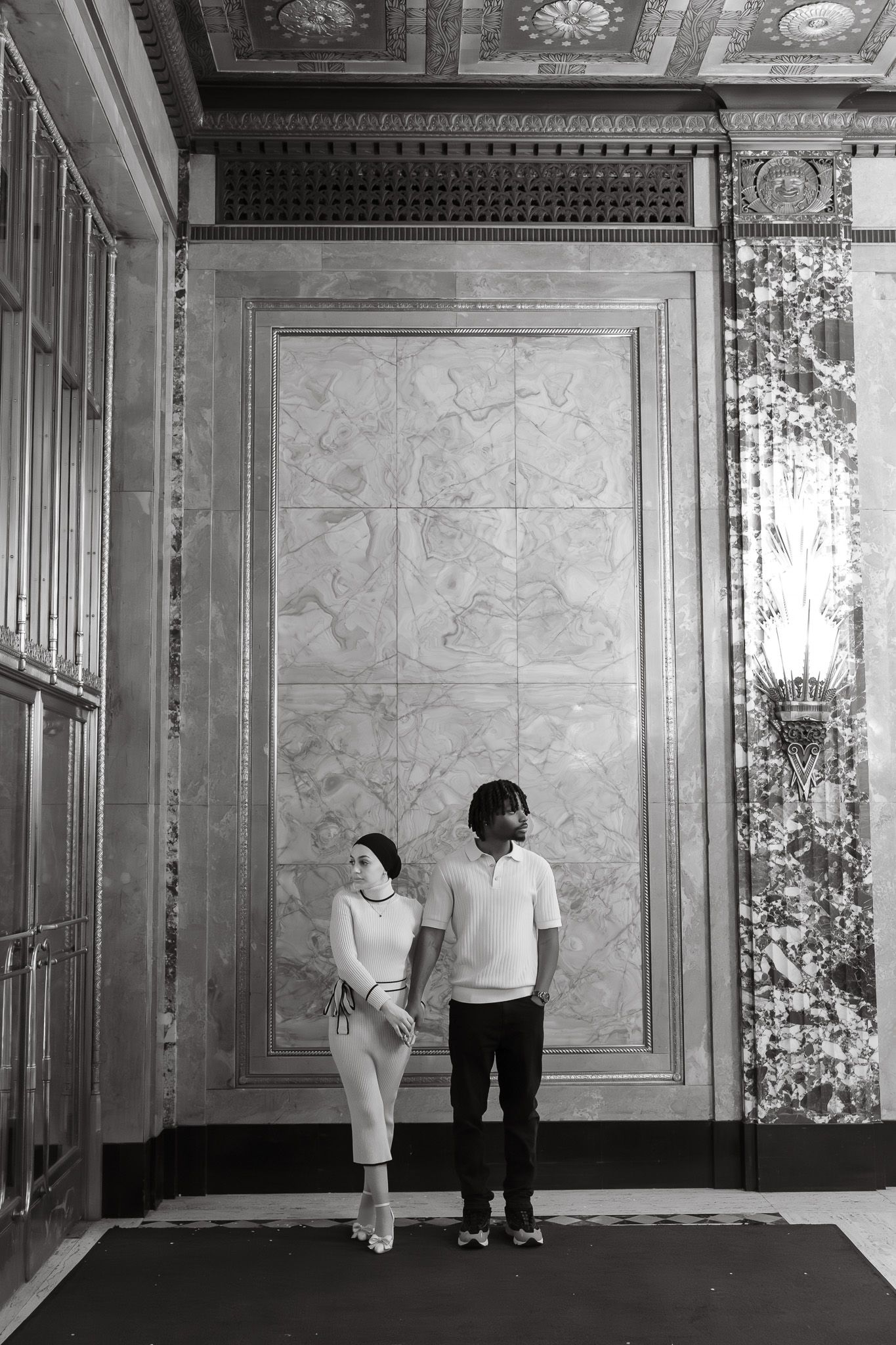Black and white photo of a couple standing in an elegant marble-walled interior with ornate ceiling details.