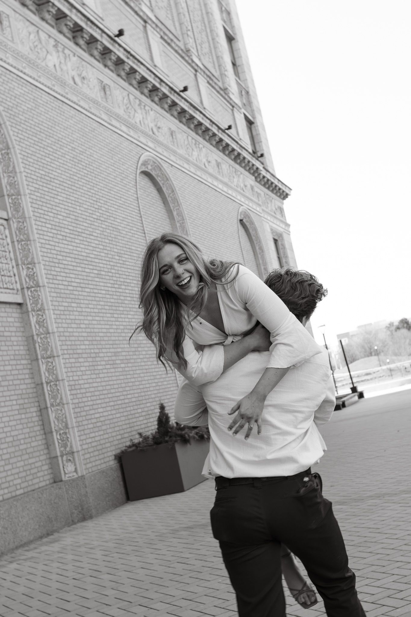 Black and white photo of a laughing woman being lifted by a man outside a brick building.