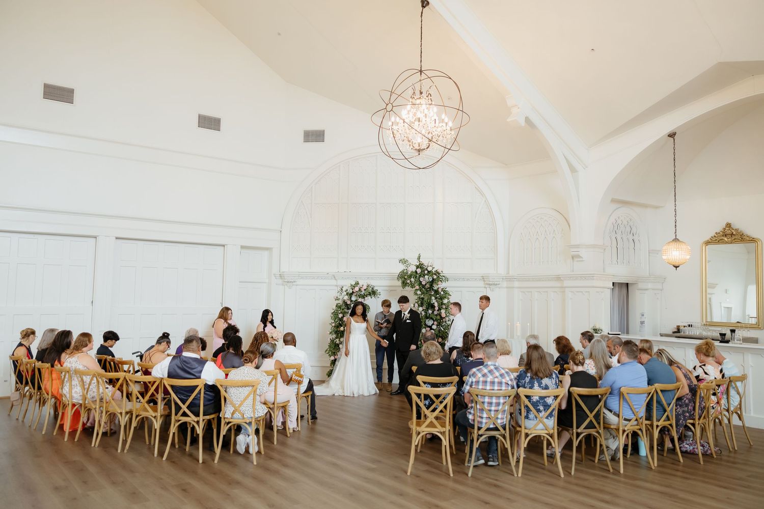 Intimate wedding ceremony in bright white venue with wooden cross-back chairs, floral arch, and elegant chandelier overhead.