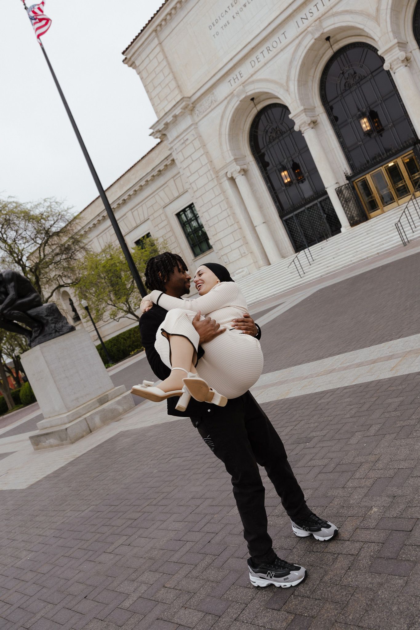 Couple sharing a romantic moment outside the Detroit Institute of Arts, man lifting woman in a loving embrace.
