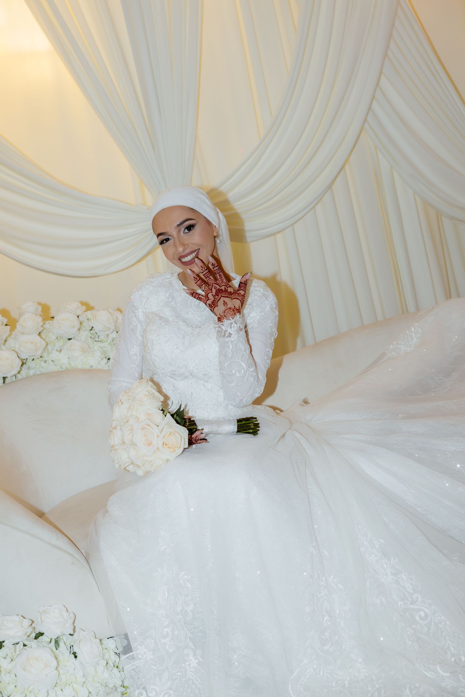 Muslim bride in white hijab and lace wedding gown with henna hands holding bouquet at elegant ceremony.