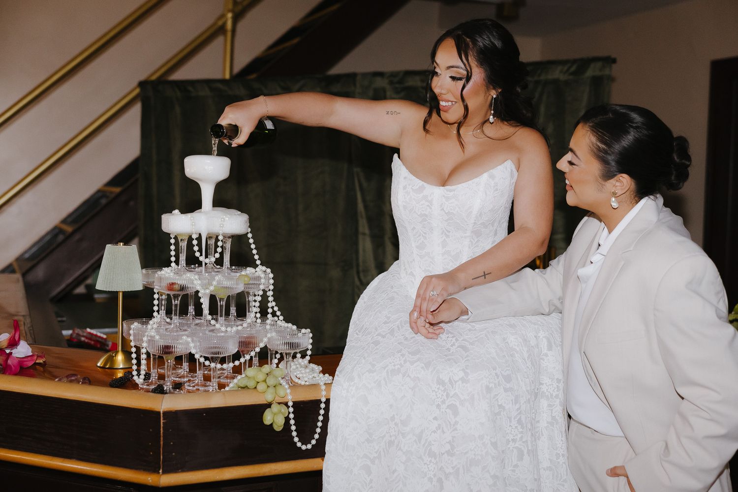 Bride in strapless gown pouring champagne tower at wedding reception while guest watches admiringly.