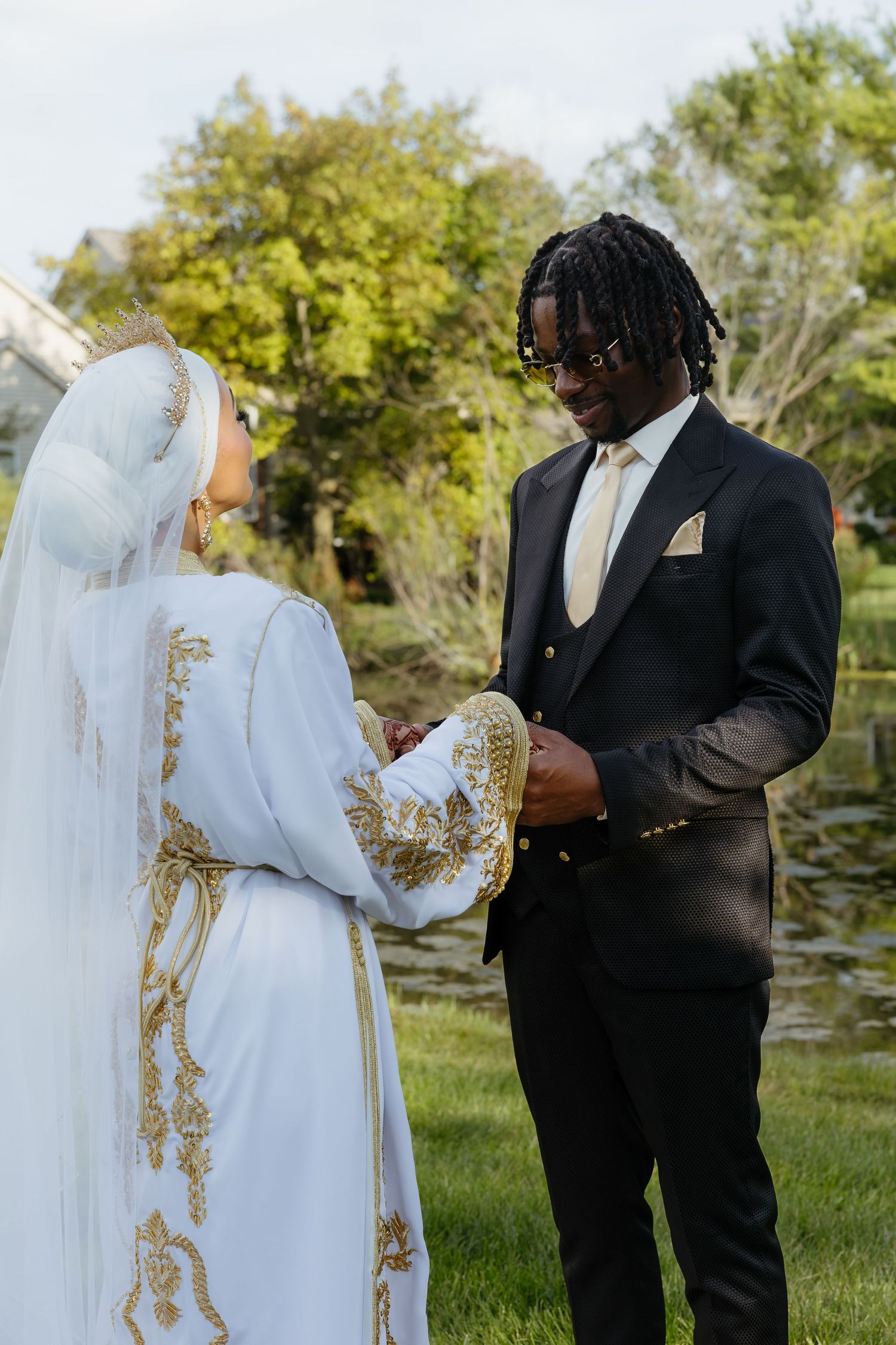 Interracial couple exchanging vows outdoors, bride in white and gold embroidered gown with veil, groom in black suit.