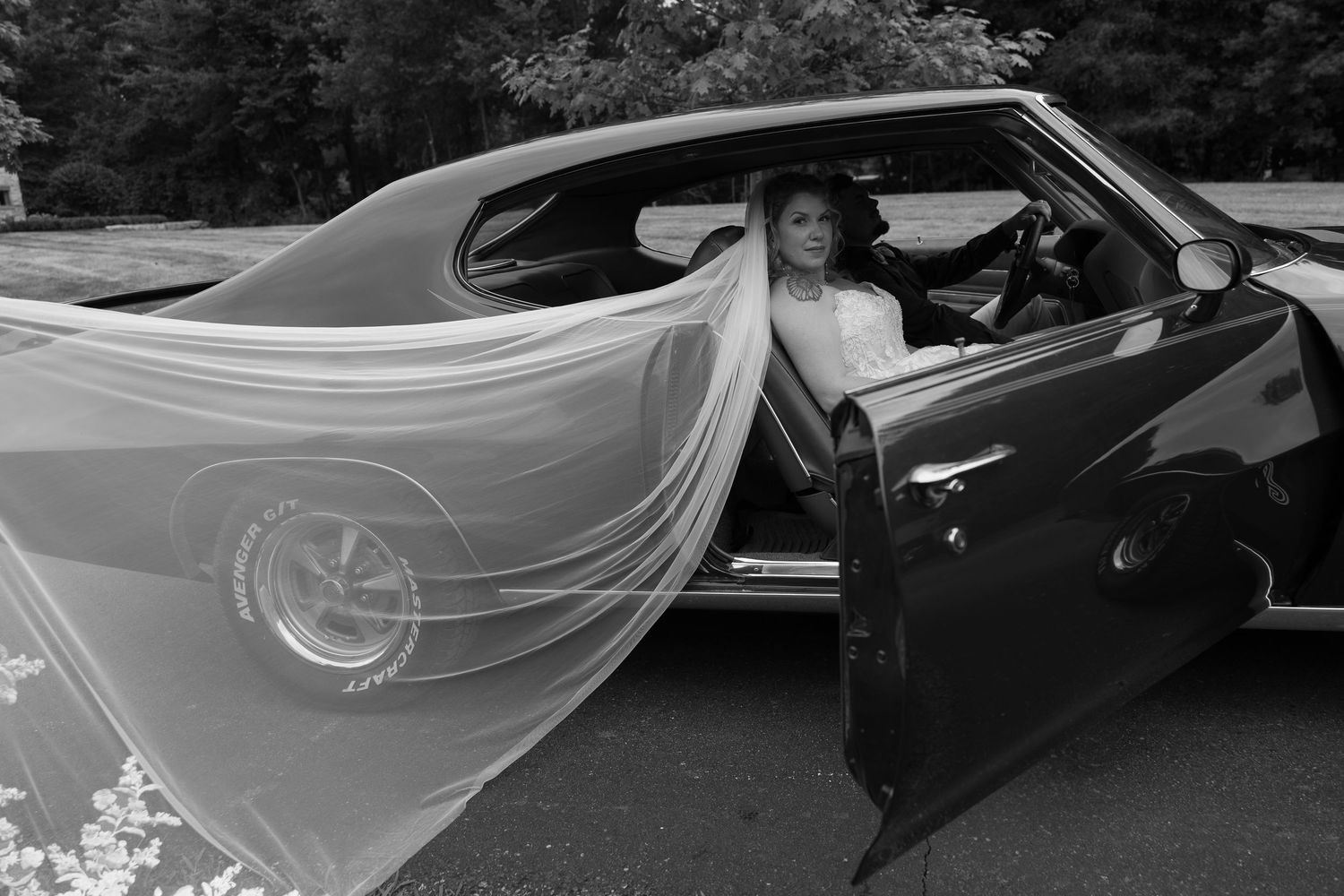 A flowing white wedding veil catches the breeze next to a vintage automobile.