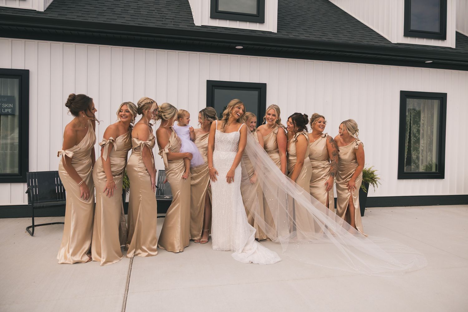 Wedding party in champagne-colored dresses poses playfully in front of white building with black trim.