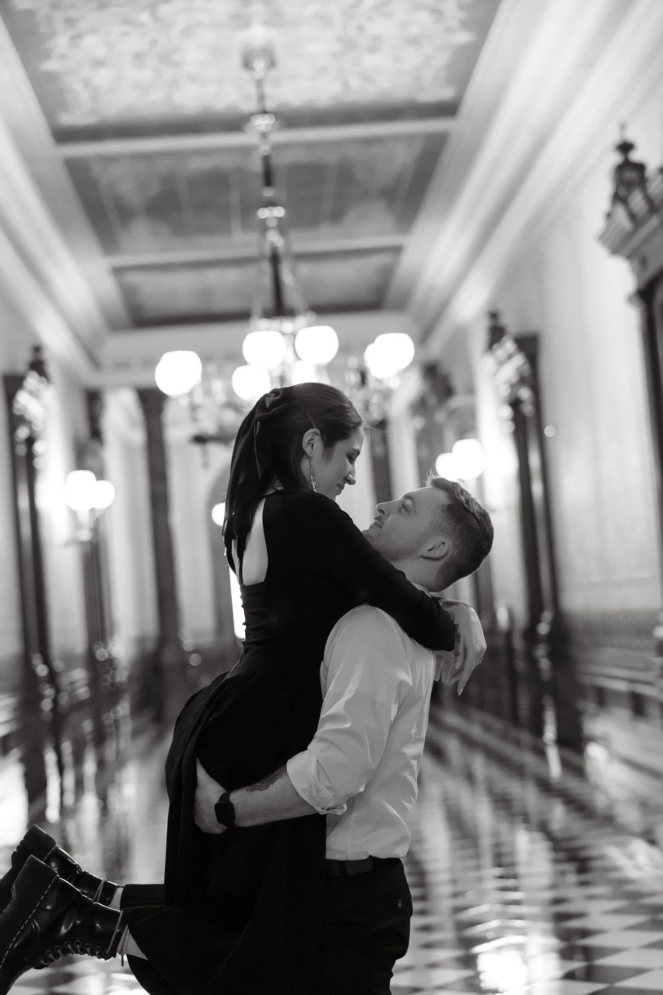 Black and white photo of a couple embracing romantically in an elegant chandelier-lit hallway.