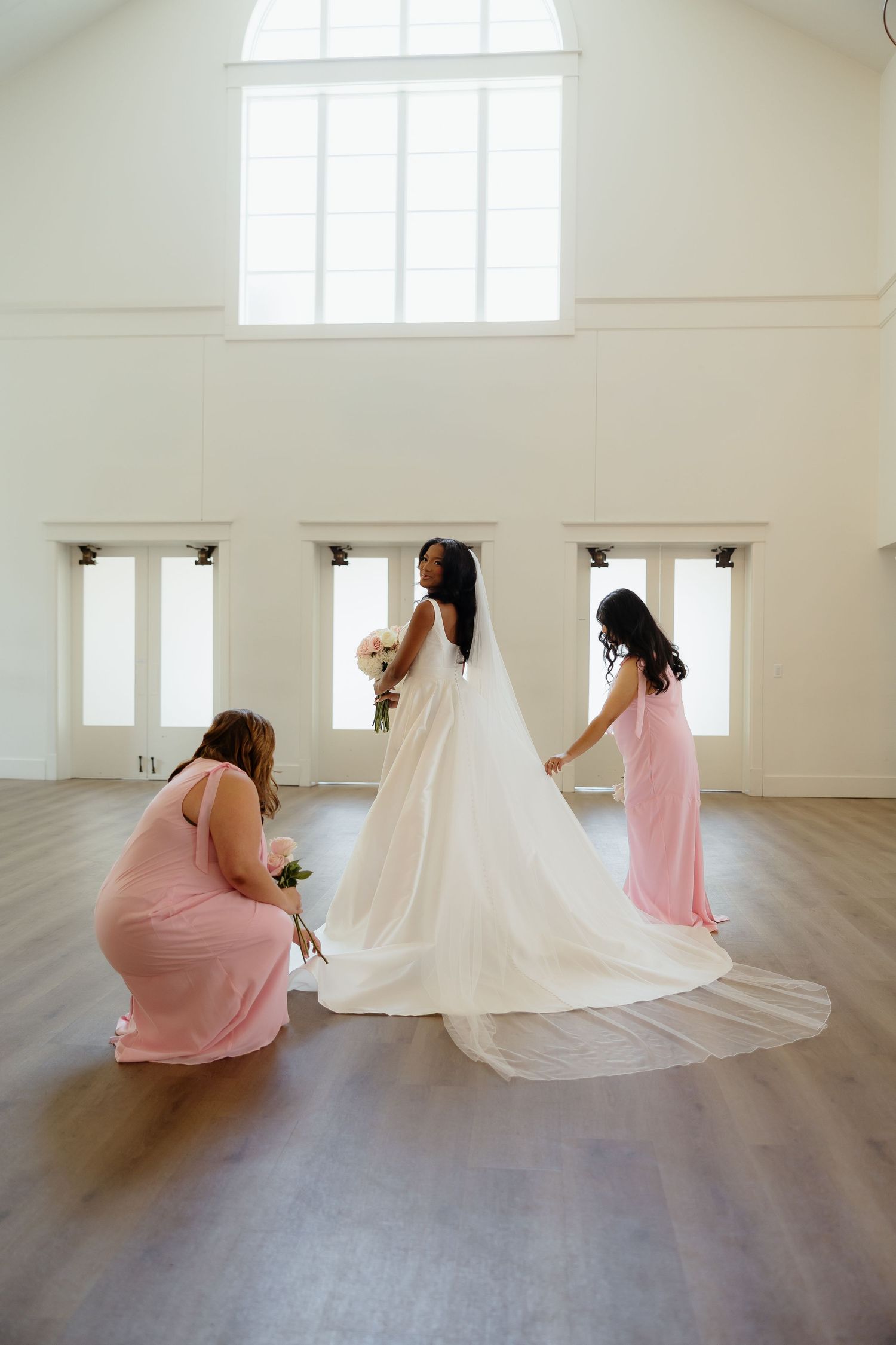 Bride in white gown holds bouquet while two bridesmaids in pink dresses arrange her train in bright white venue.