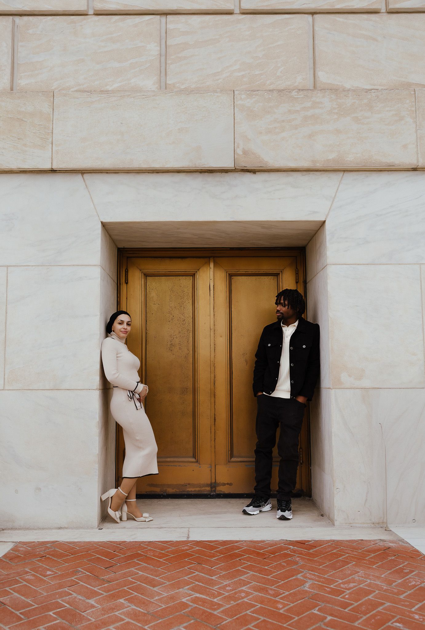 Couple posing in front of a wooden door set in a marble wall, woman in white dress and man in black jacket.