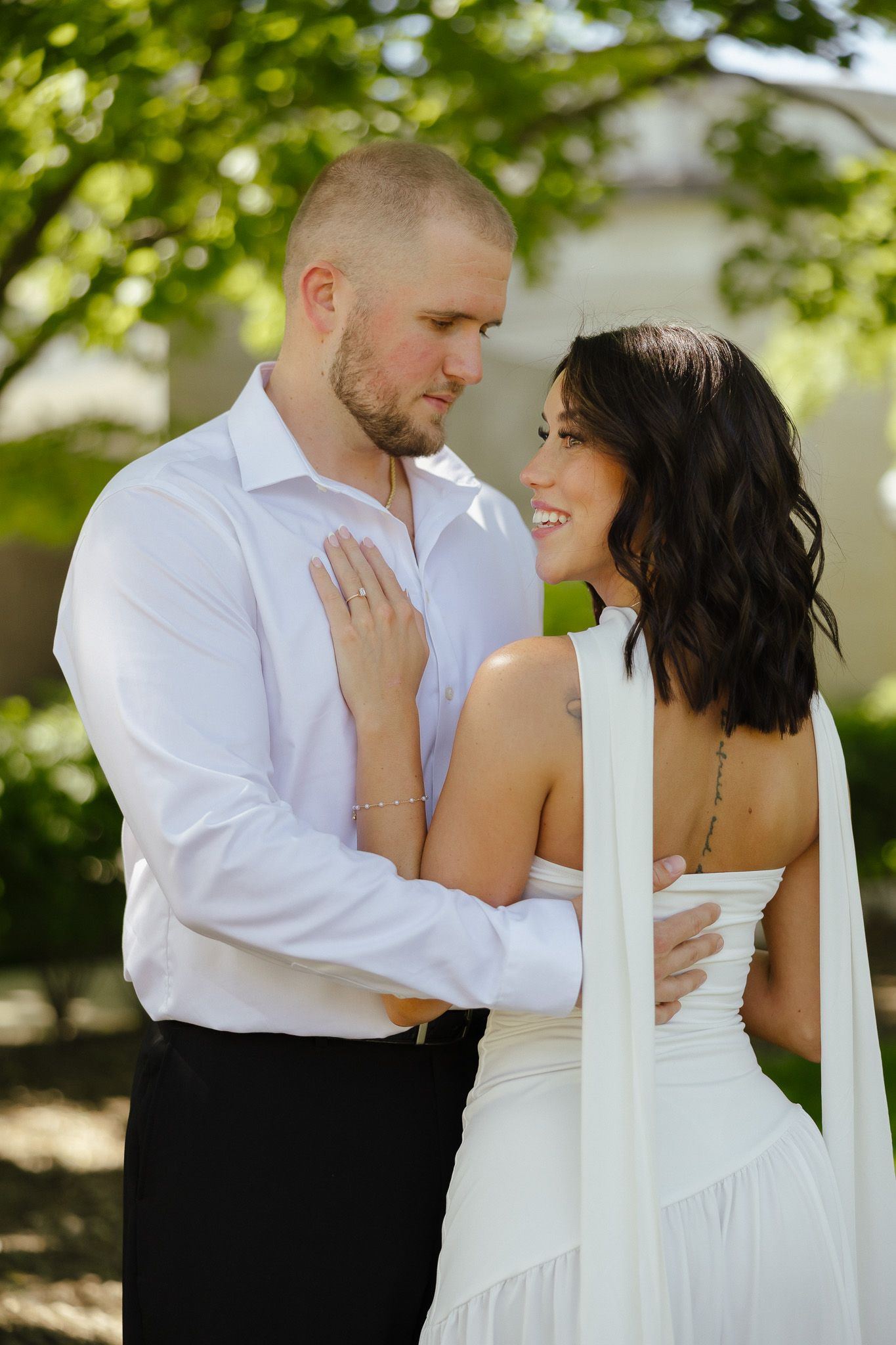 A couple in white wedding attire embrace outdoors, smiling warmly beneath lush green trees in soft natural light.