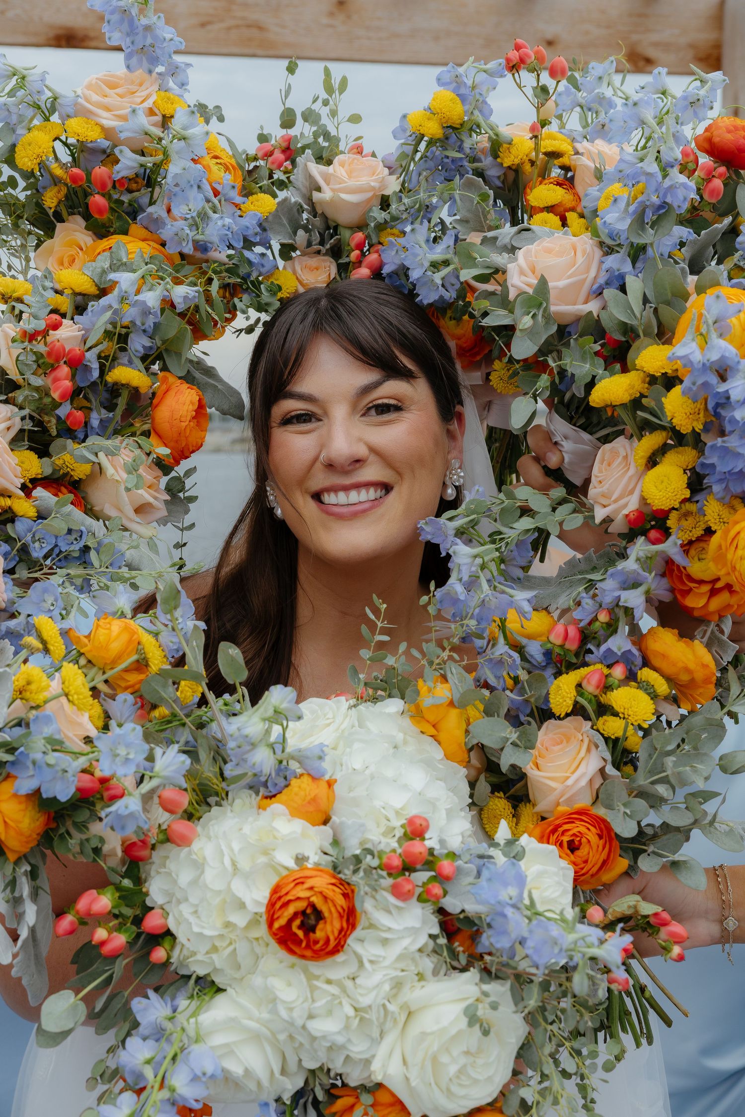 Smiling bride holding white and orange bouquet framed by colorful floral arrangement with blue, yellow, and orange blooms.