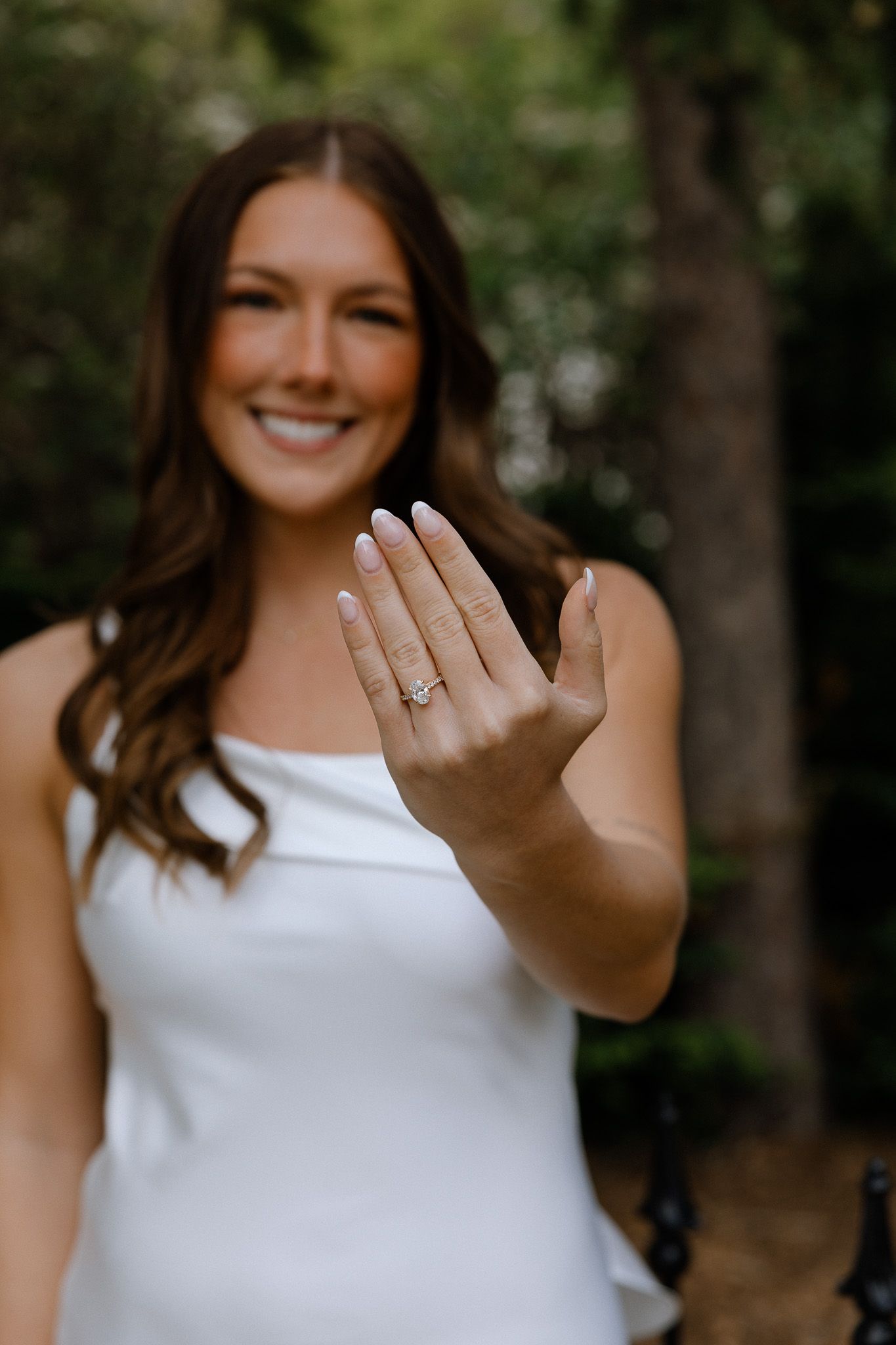 Radiant bride in white dress proudly displays her engagement ring toward the camera outdoors.
