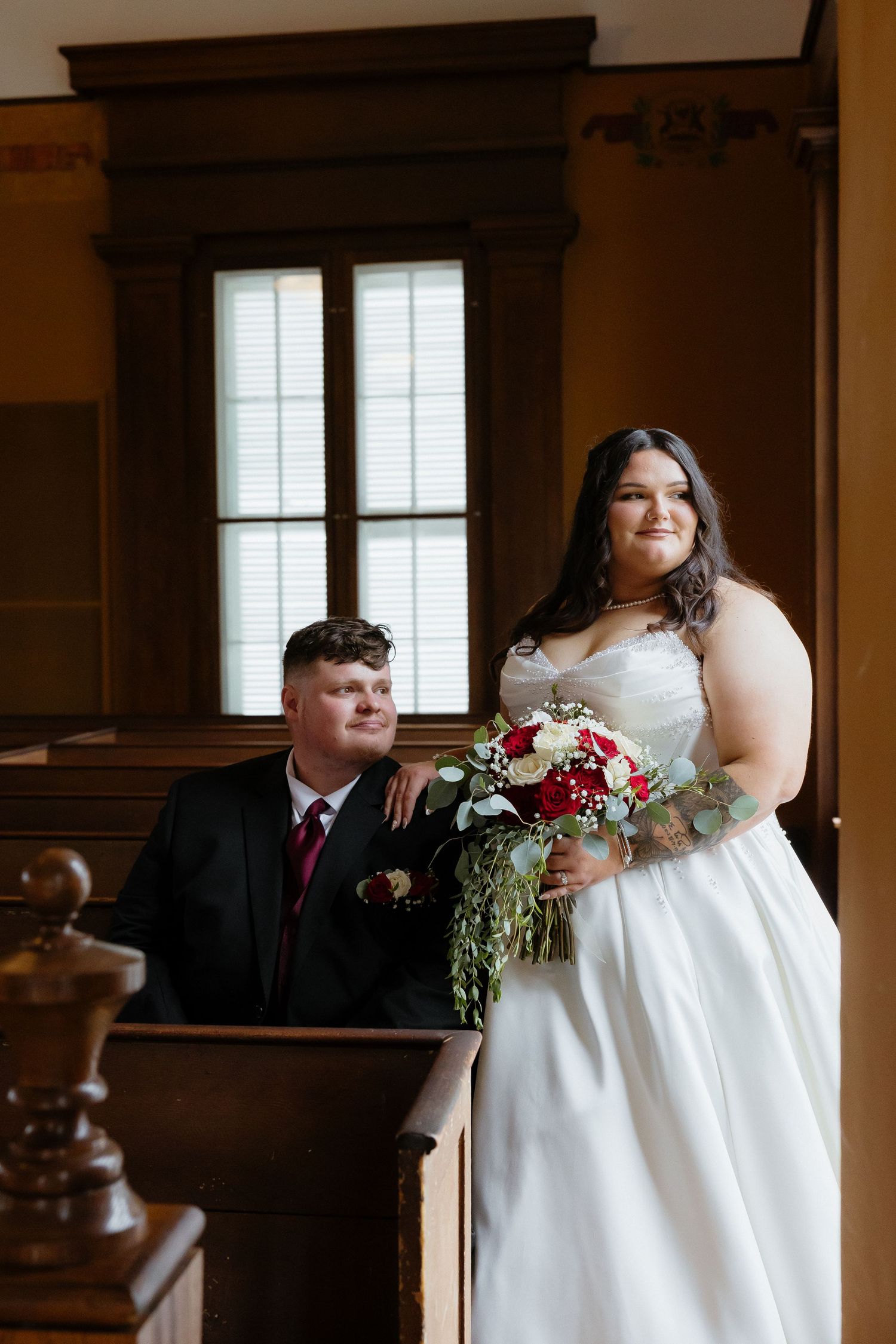 Bride in white gown holding red and white bouquet stands beside groom in black suit inside a wooden church.