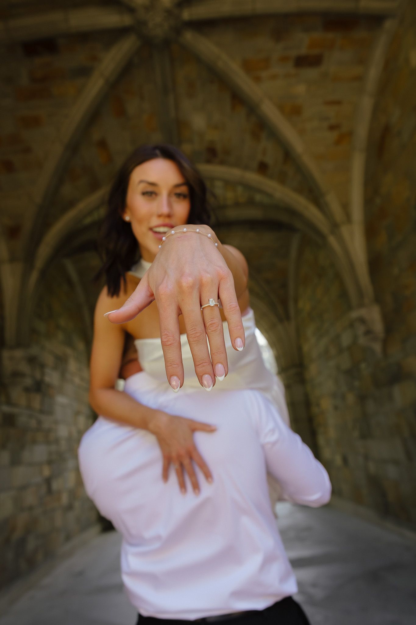 Bride shows off engagement ring while being carried by groom in historic stone archway venue.