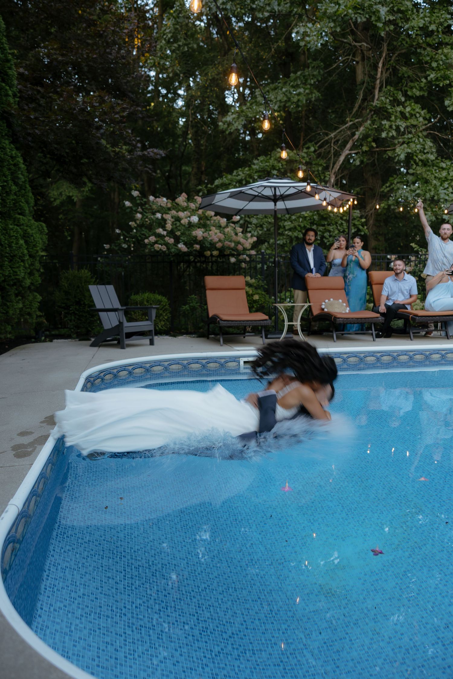 Bride in white wedding gown jumping into backyard pool while wedding guests cheer and celebrate poolside at dusk.