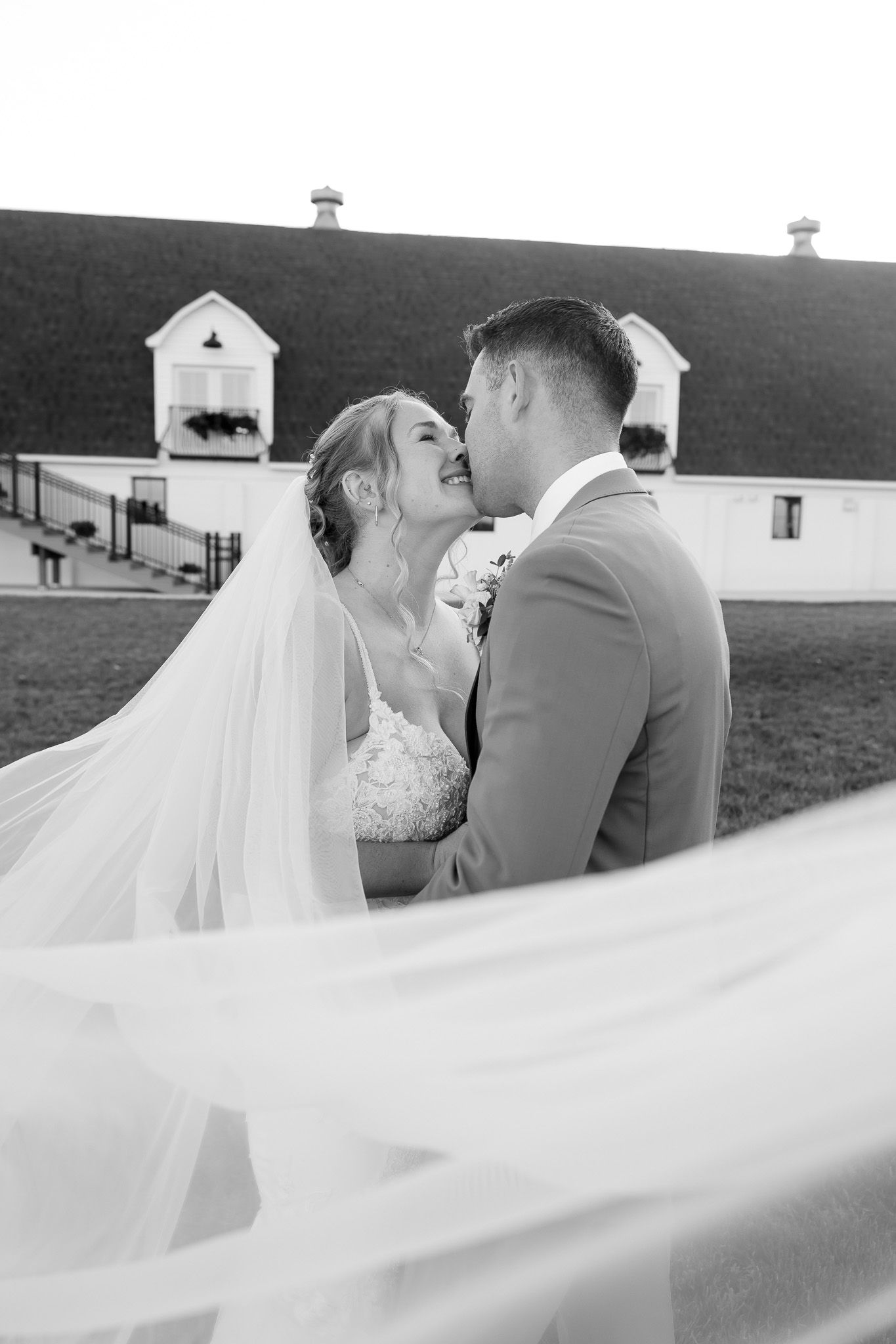 Black and white photo of a bride and groom sharing a romantic moment, with a white barn in the background.