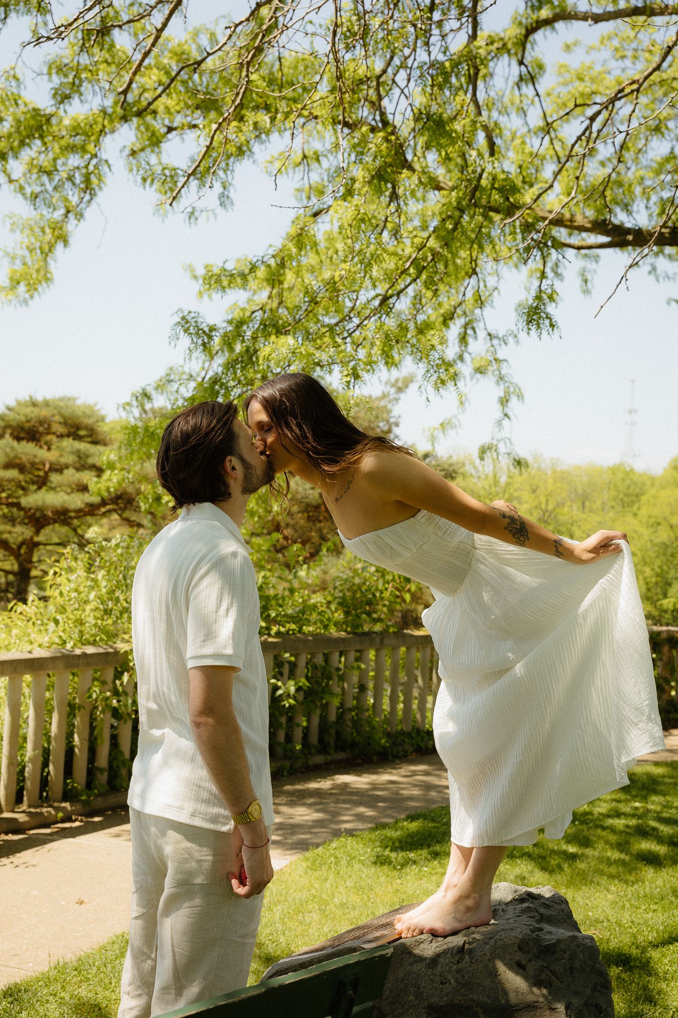A couple shares a romantic kiss outdoors, woman in white dress standing on rock, man in white outfit, lush green park setting.