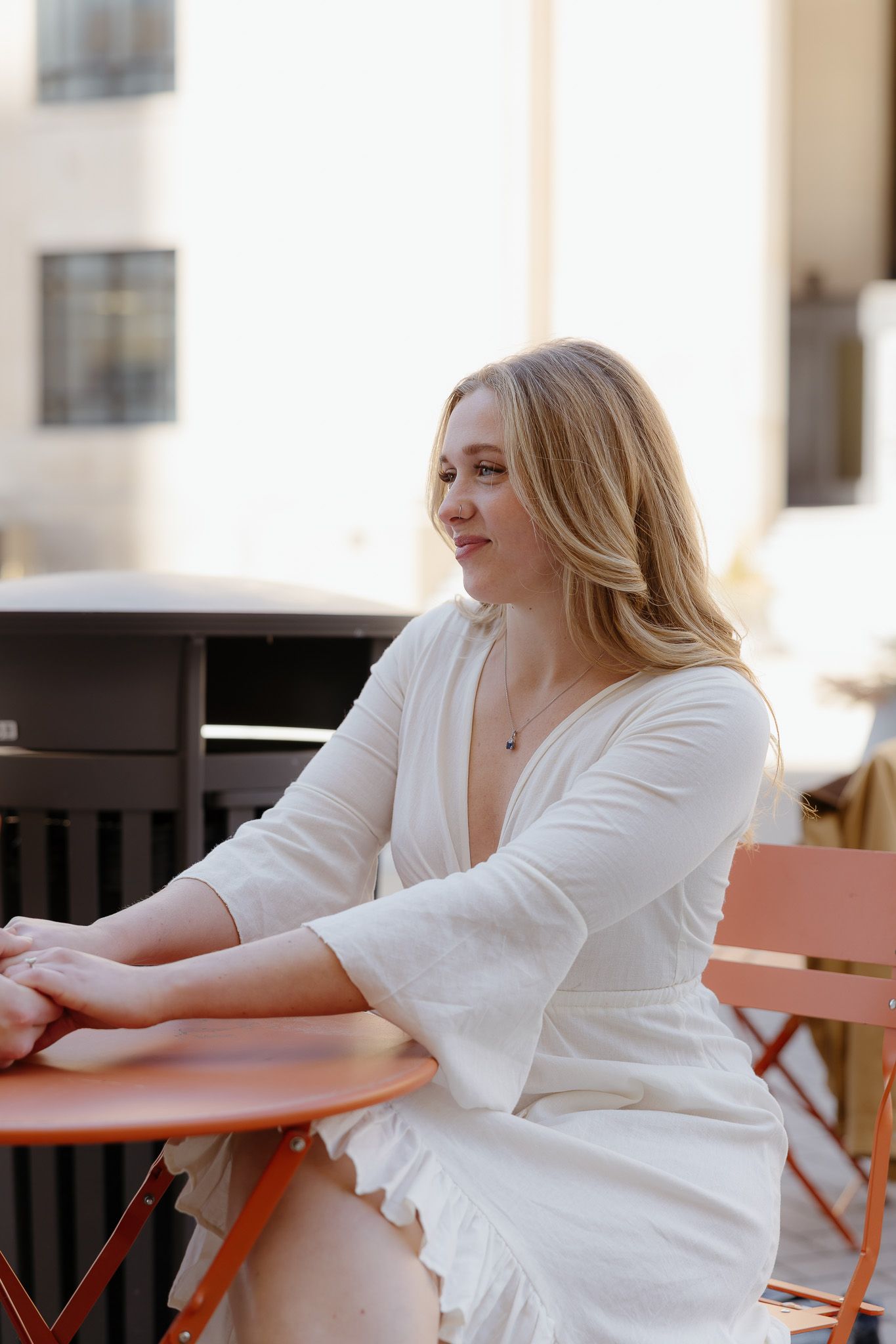 Blonde woman in white dress sitting at orange outdoor cafe table, gazing away in urban setting.