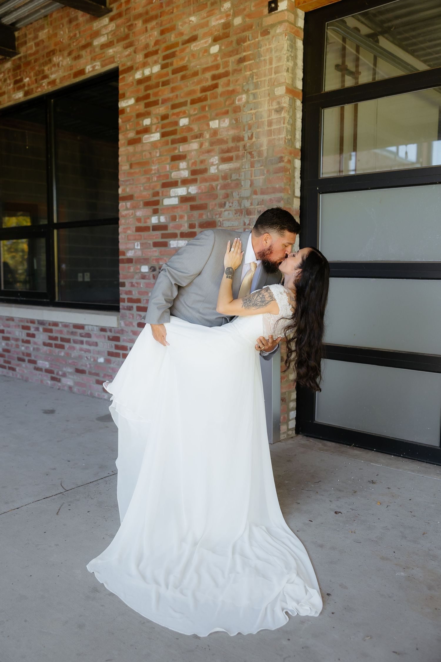 A groom dips his bride for a romantic kiss outside a rustic brick building on their wedding day.