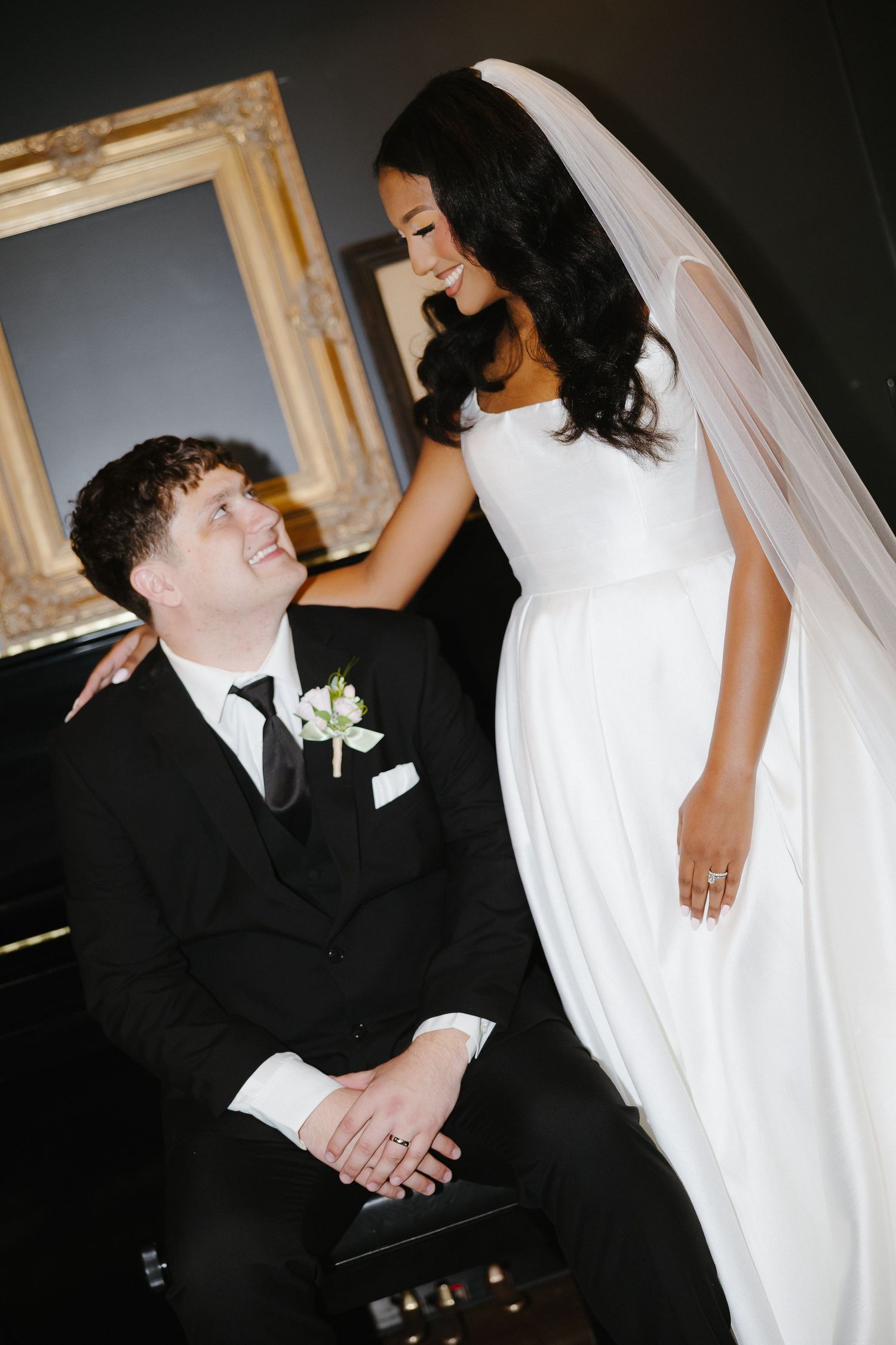 Bride in white gown and veil smiles at groom in black tuxedo seated before ornate gold frame.
