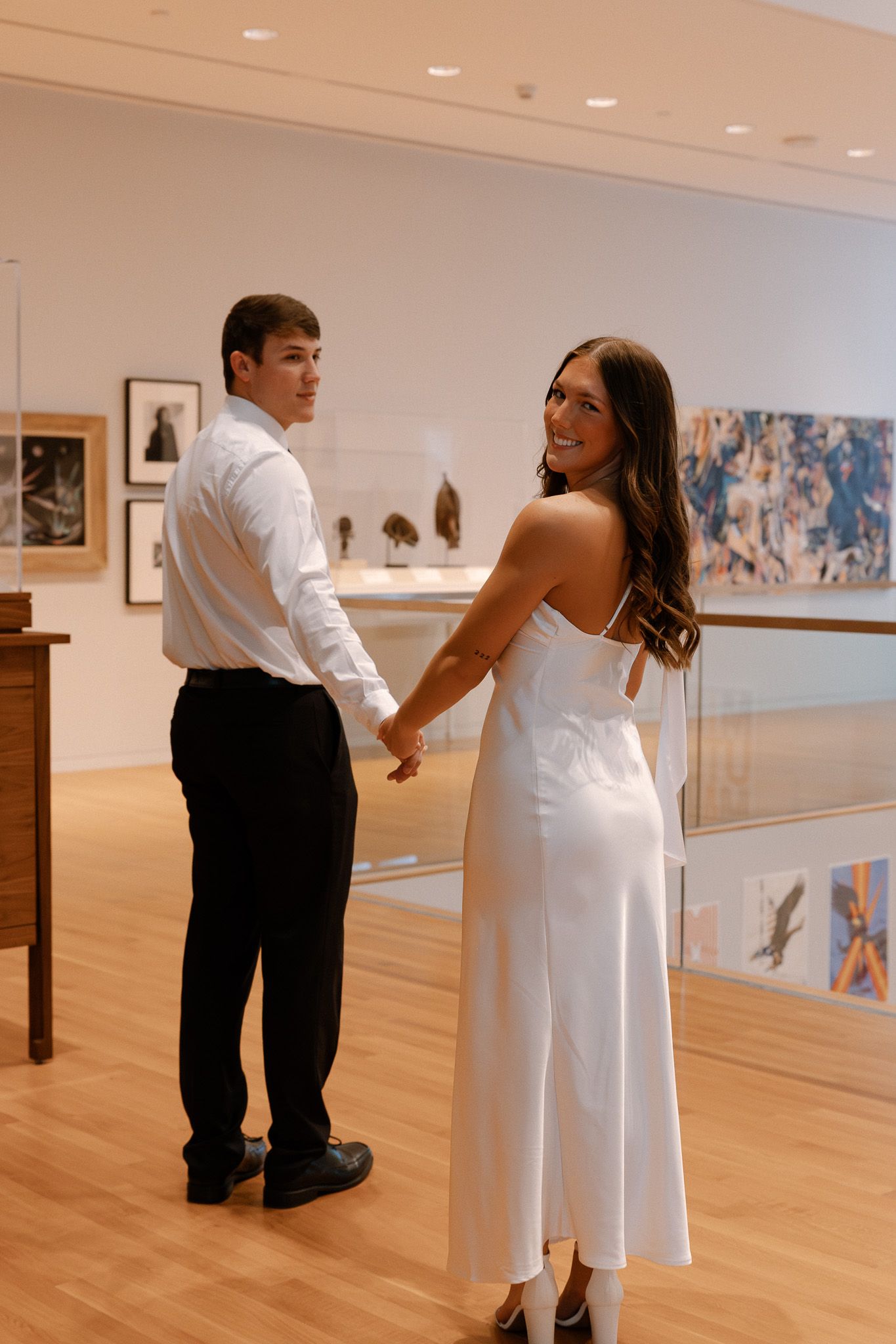 Couple holding hands in white dress and black pants posing in an art museum gallery with paintings on walls.