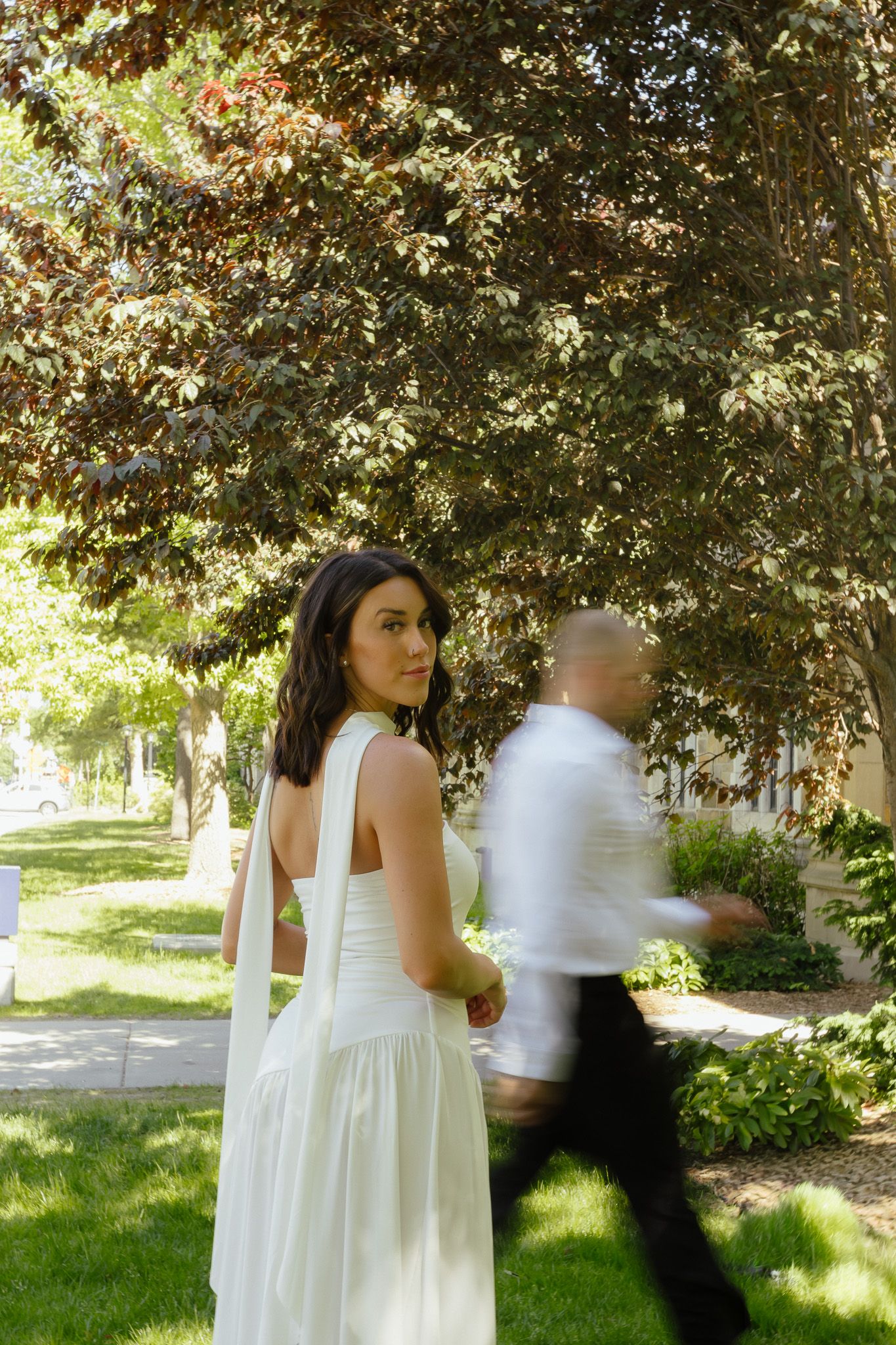 Bride in white dress looks back while groom walks past in sunny garden setting.