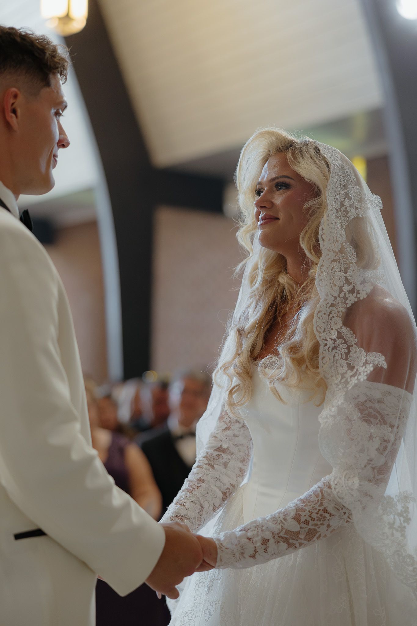 Smiling blonde bride in lace veil holds hands with groom in white tuxedo during indoor wedding ceremony.