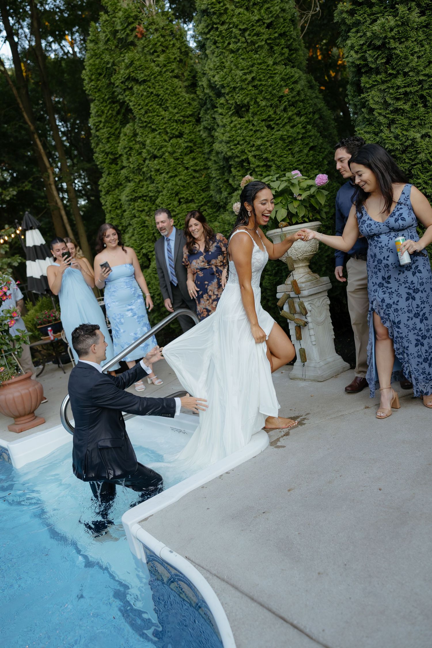 Bride in white gown laughs as groom sits in pool at outdoor wedding reception with guests watching.