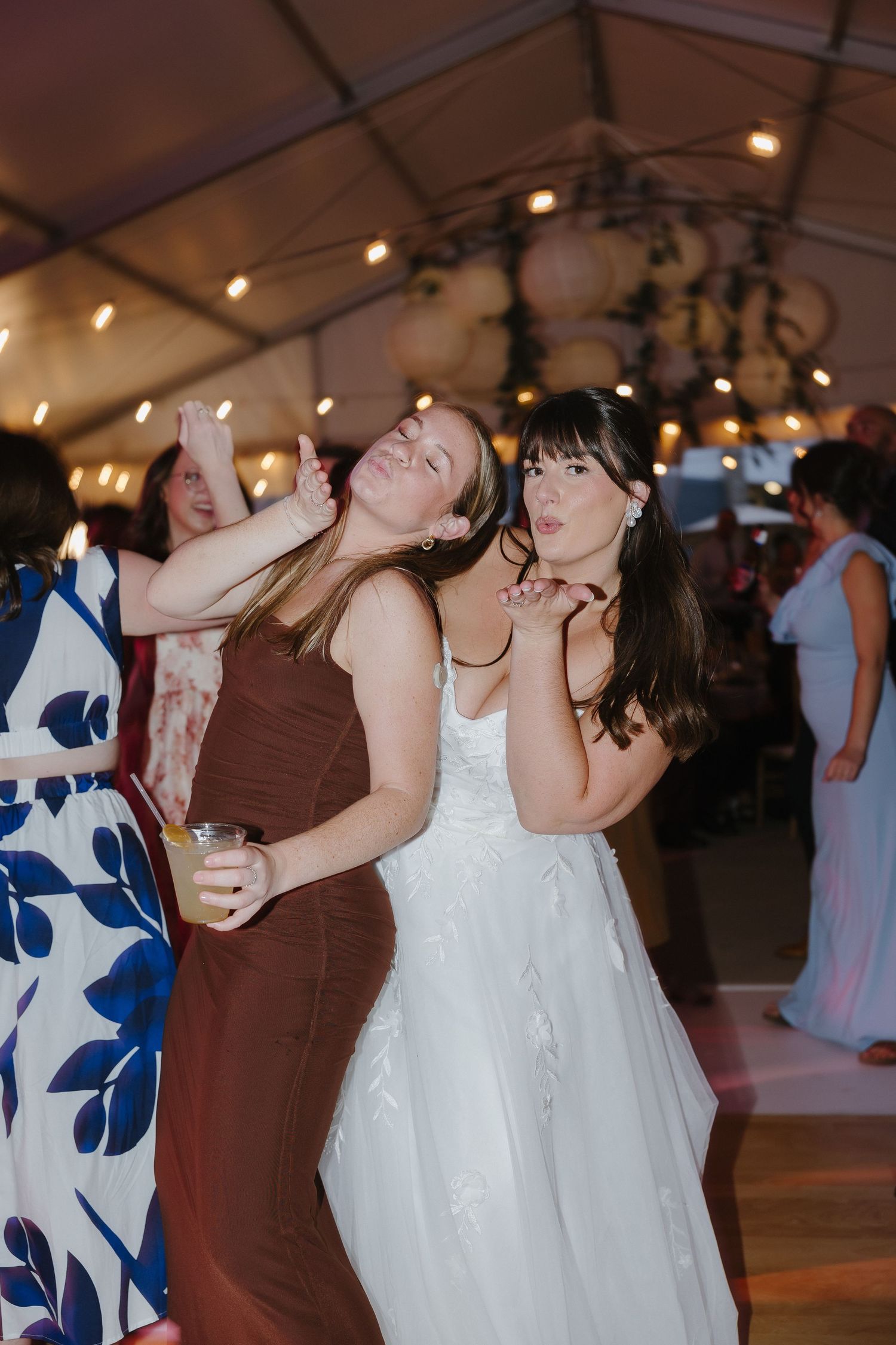 Bride in white gown poses with friend on wedding reception dance floor under warm string lights in tent.