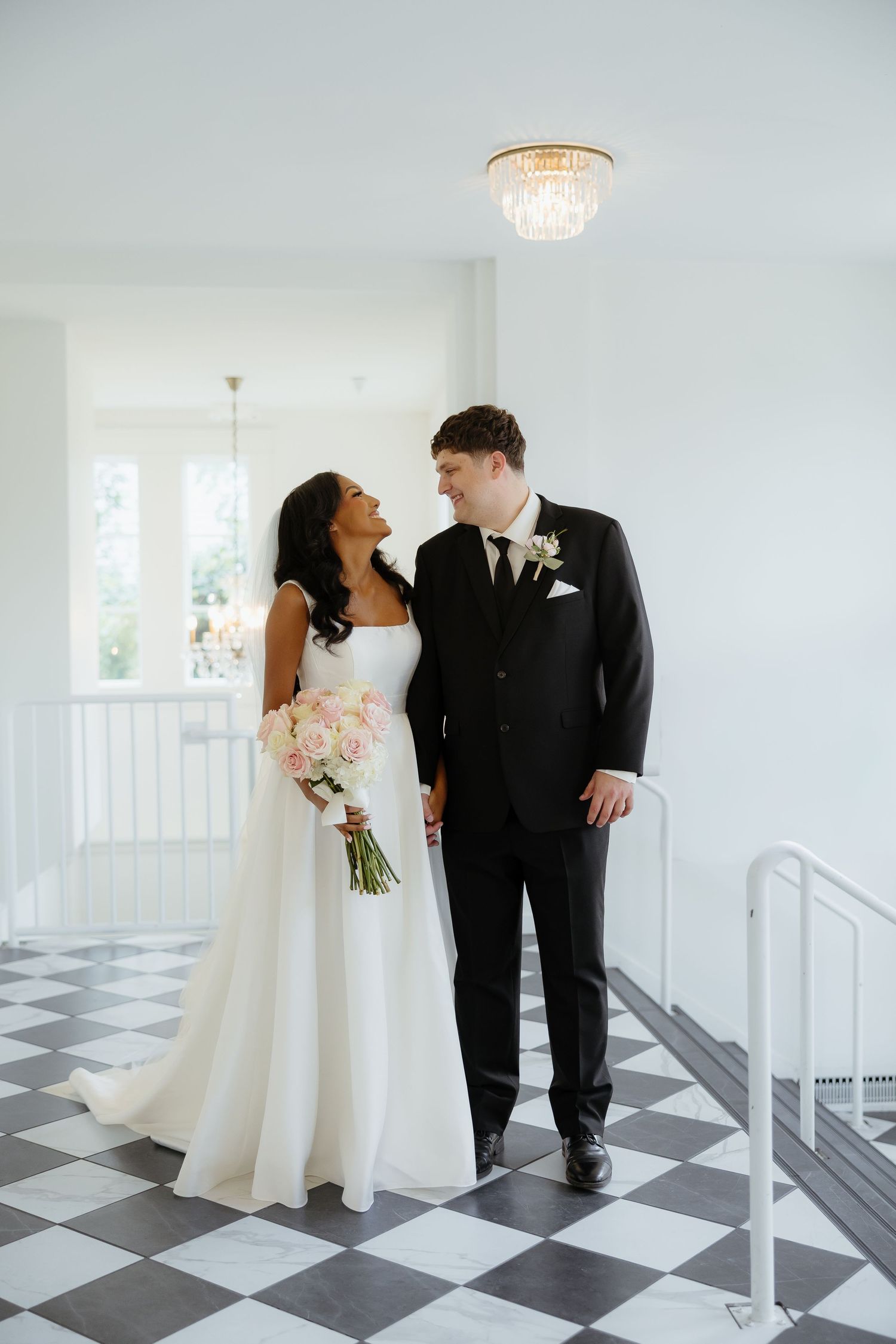Bride and groom share a tender moment on black and white checkered floor beneath a crystal chandelier.