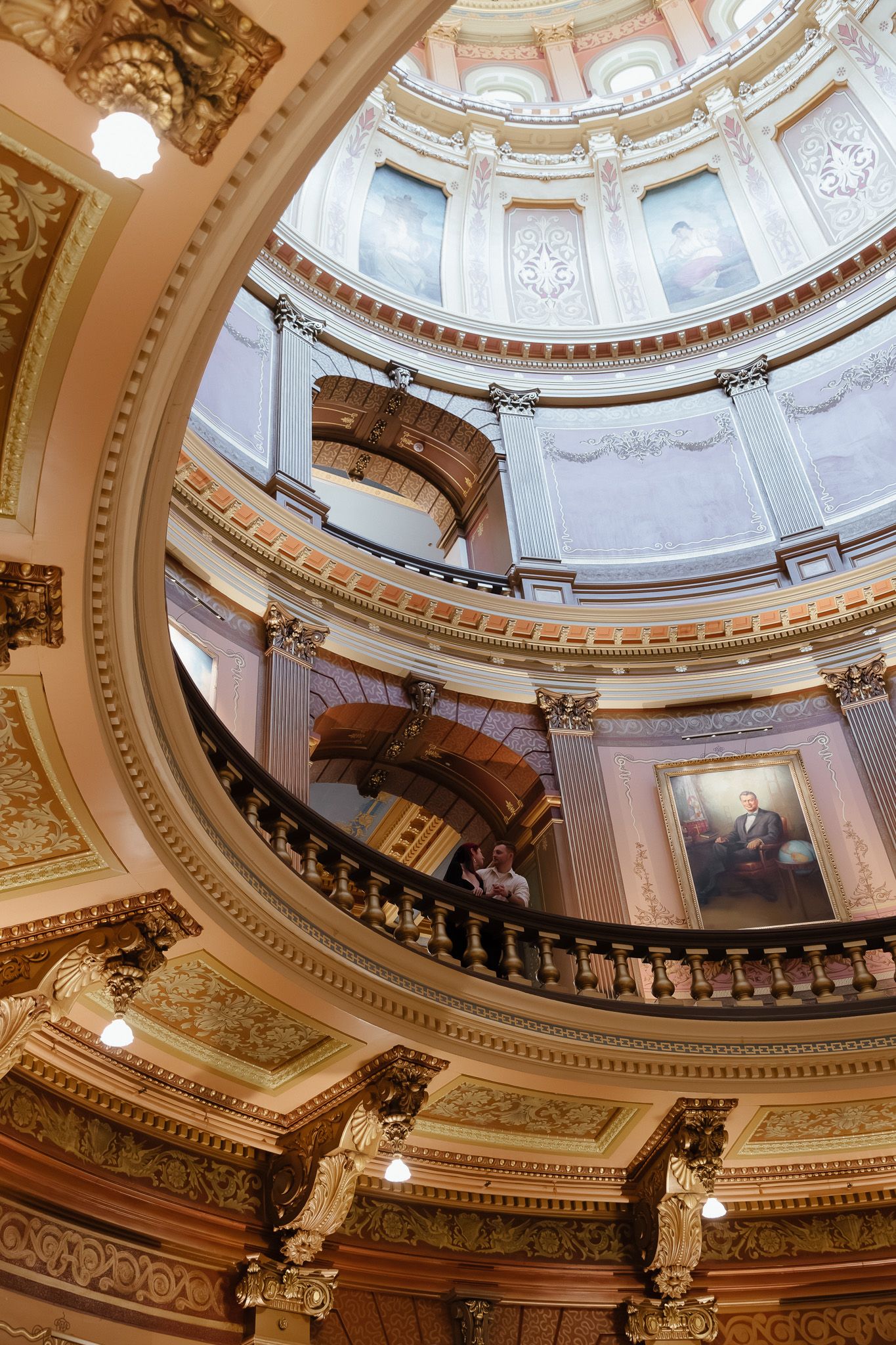 Interior view of ornate capitol building rotunda with gilded balconies, murals, and portrait painting under a grand dome.