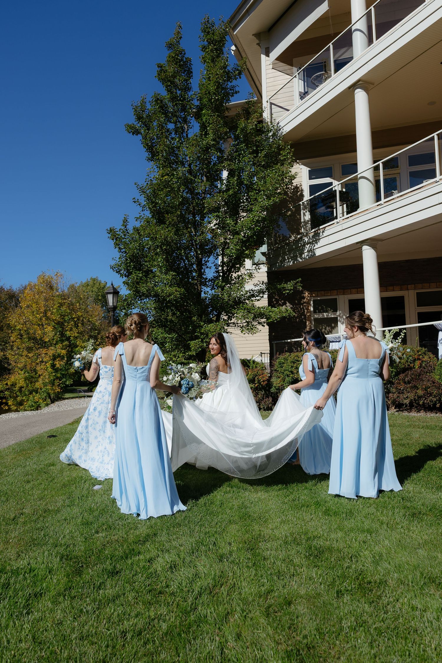 Bridesmaids in light blue gowns help carry a bride's white train across green lawn outside elegant venue on sunny day.